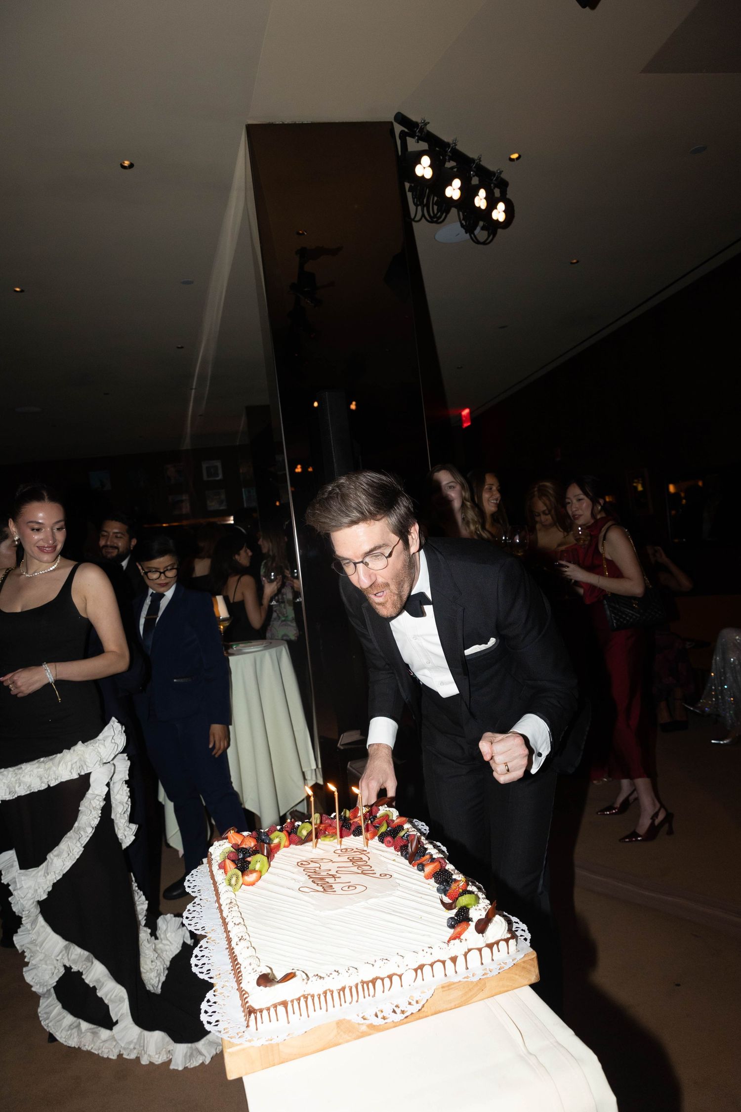 Man in a tuxedo and glasses leans over a decorated birthday cake with lit candles at a formal party as guests mingle behind him.