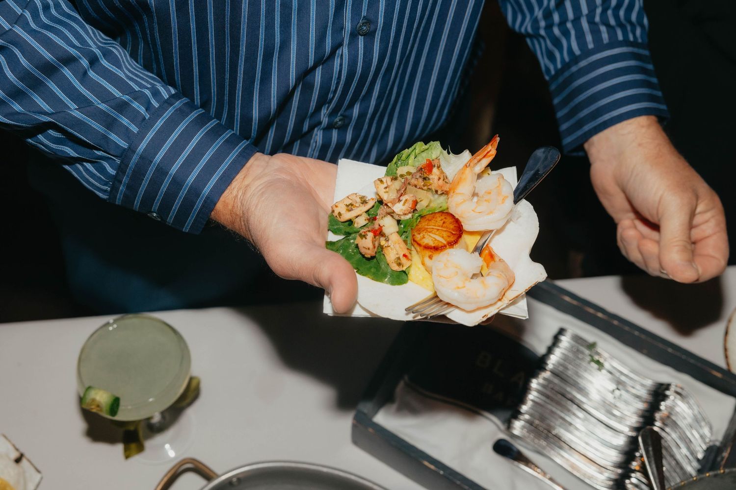 Person in a blue striped shirt holds a serving of shrimp, orange slices and fresh greens on a paper plate at a catered event.