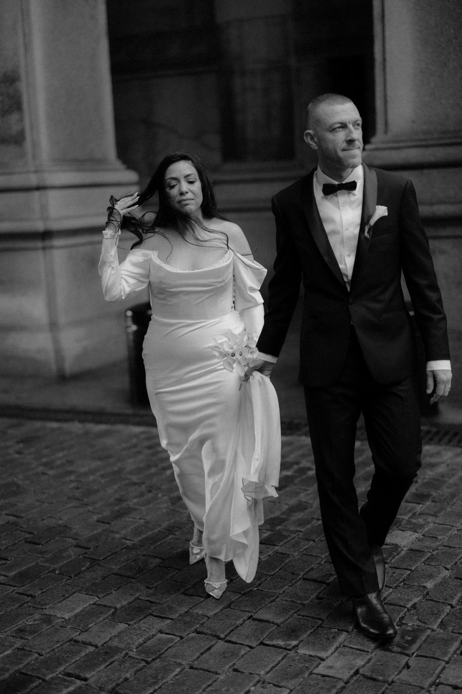 Bride and groom walk together on cobblestone street in black and white wedding photo.