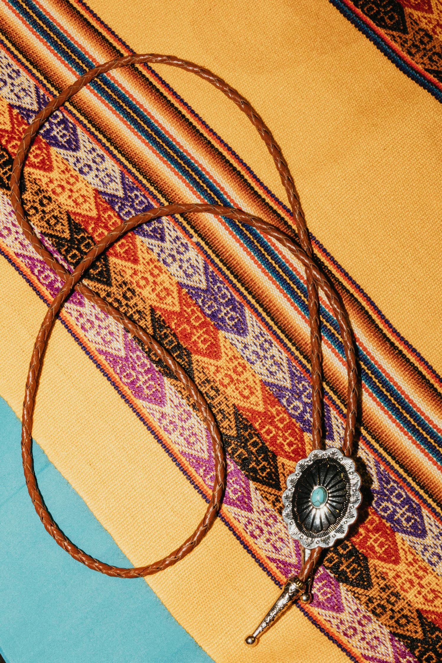 A turquoise bolo tie with silver concho rests on a colorful Southwest Native American woven textile blanket.