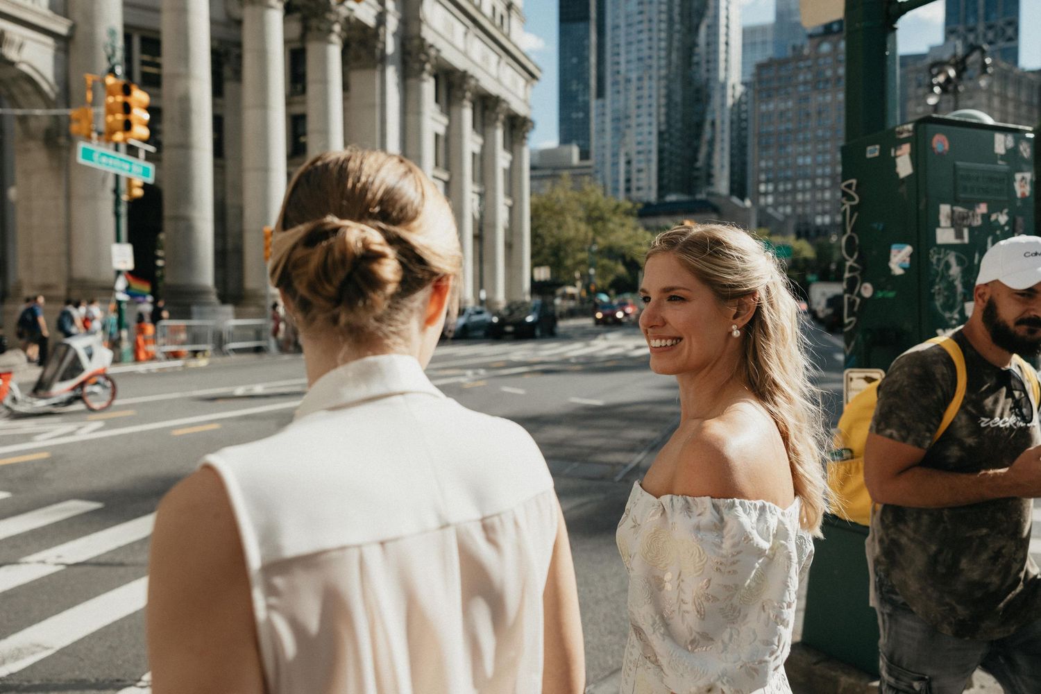 Two brides on a busy city street, one smiling in an off-shoulder floral dress on their wedding day.