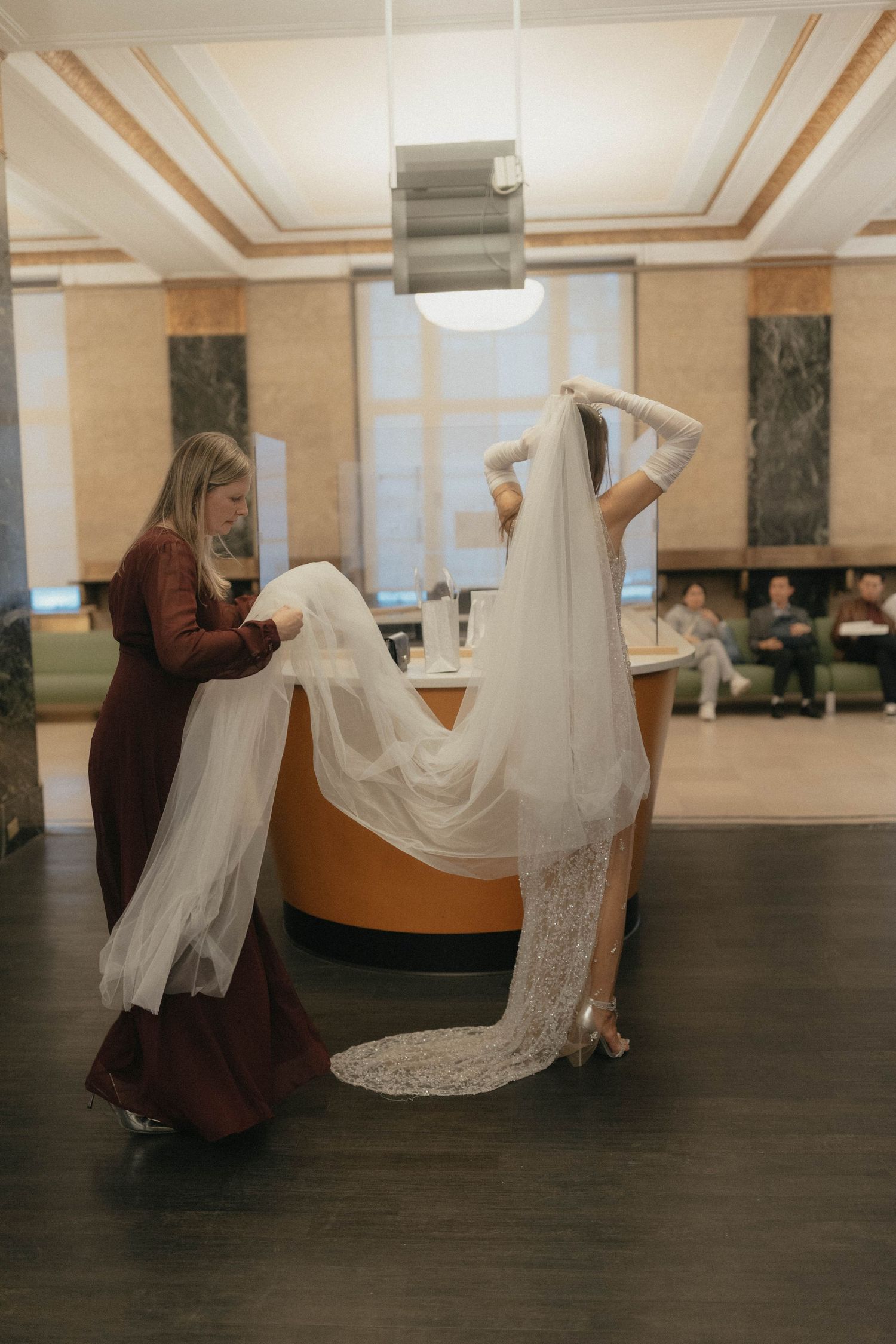 Bride adjusting long lace veil assisted by woman in brown dress in elegant lobby interior.