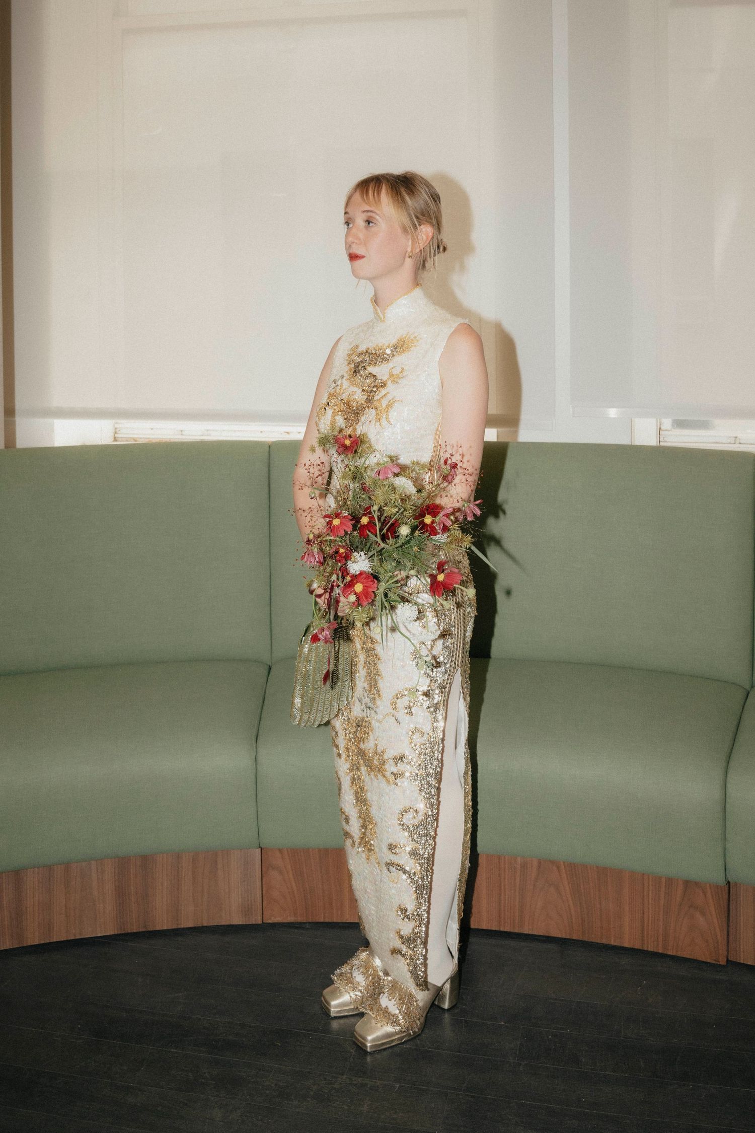 Bride in embroidered white gown holds red and green bouquet beside curved green sofa.