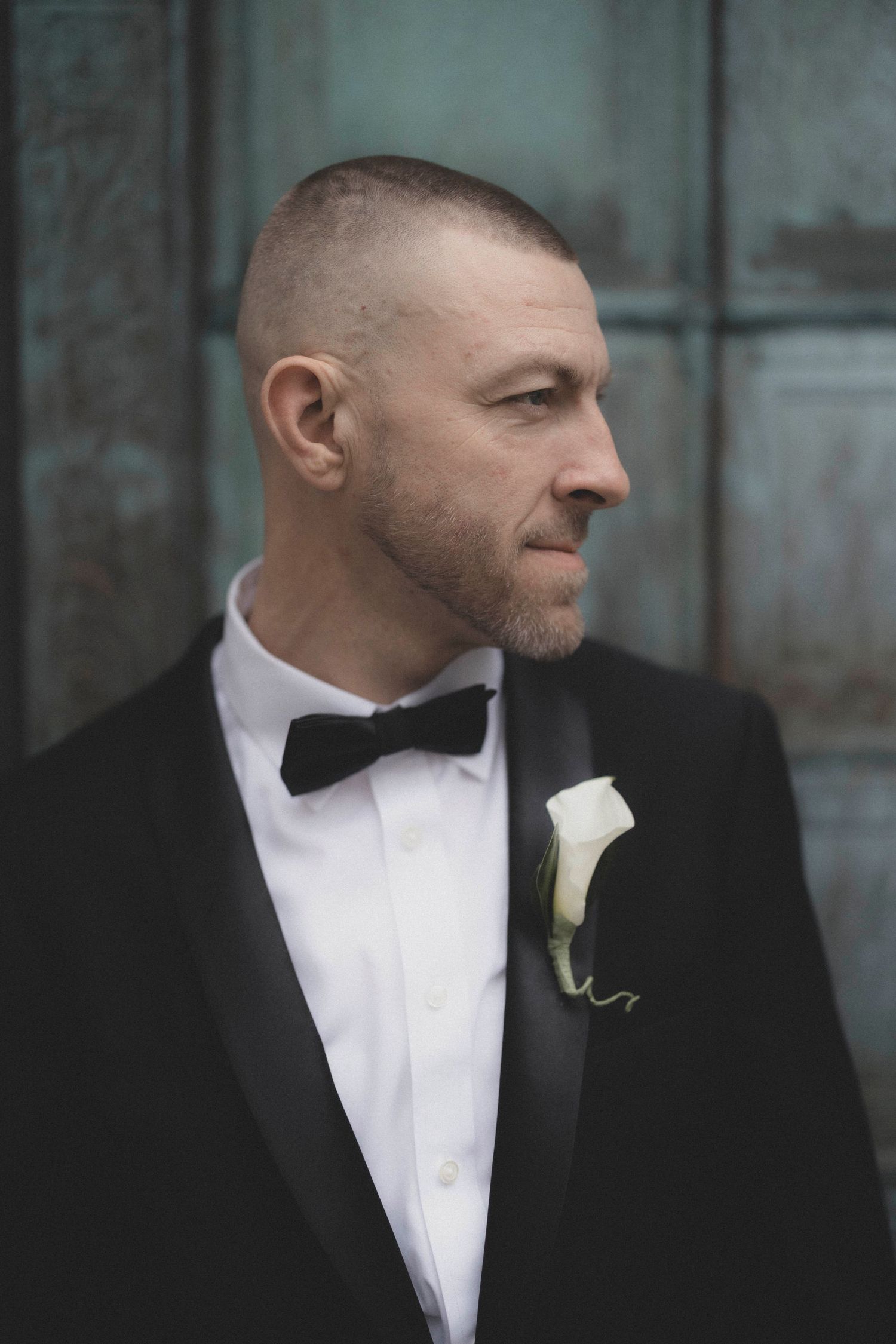 Handsome groom in black tuxedo with bow tie and white calla lily boutonniere looking away.