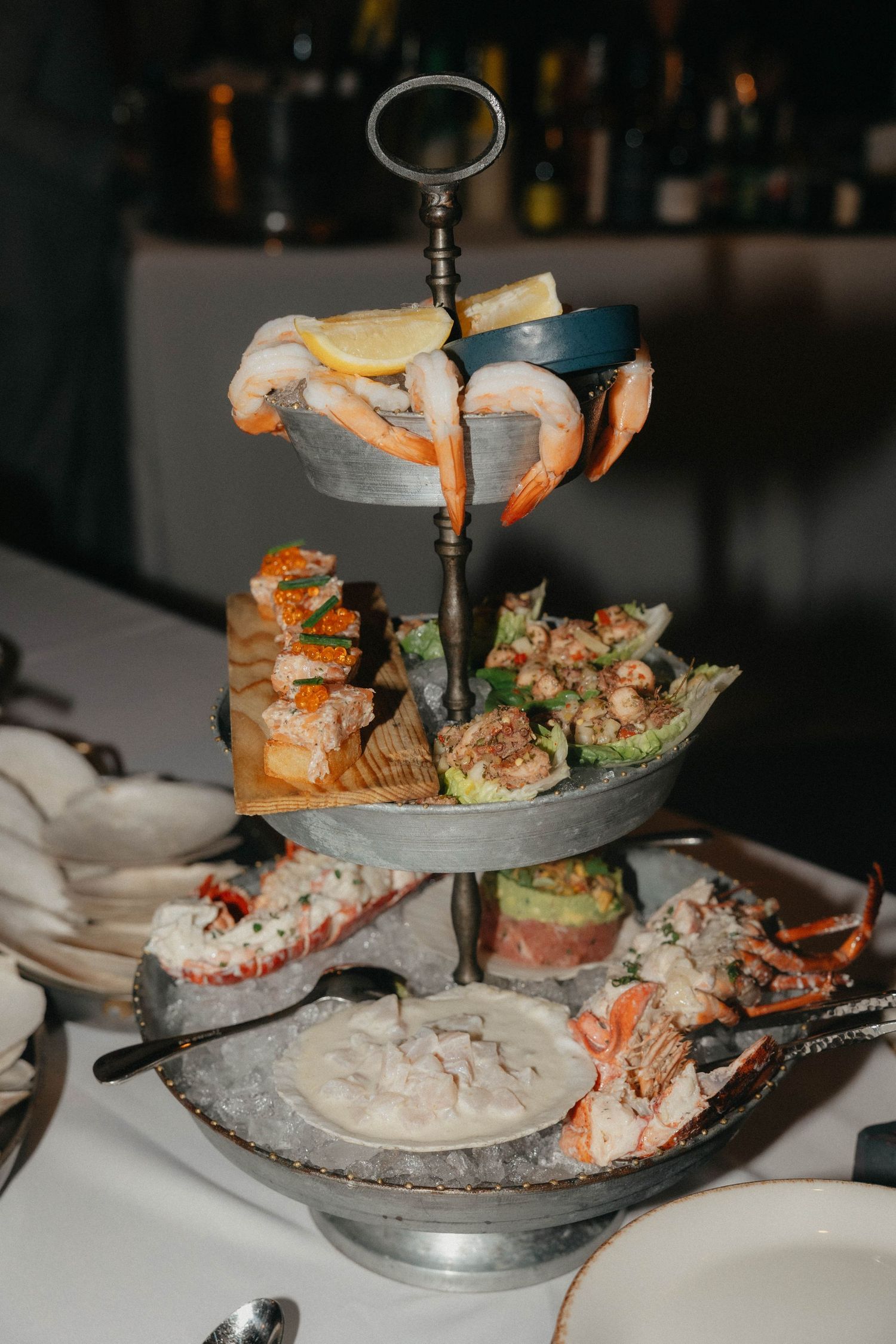 Three-tiered metal seafood tower with shrimp, lemon wedges, crab claws, lobster tails and fresh oysters on ice at an upscale restaurant.