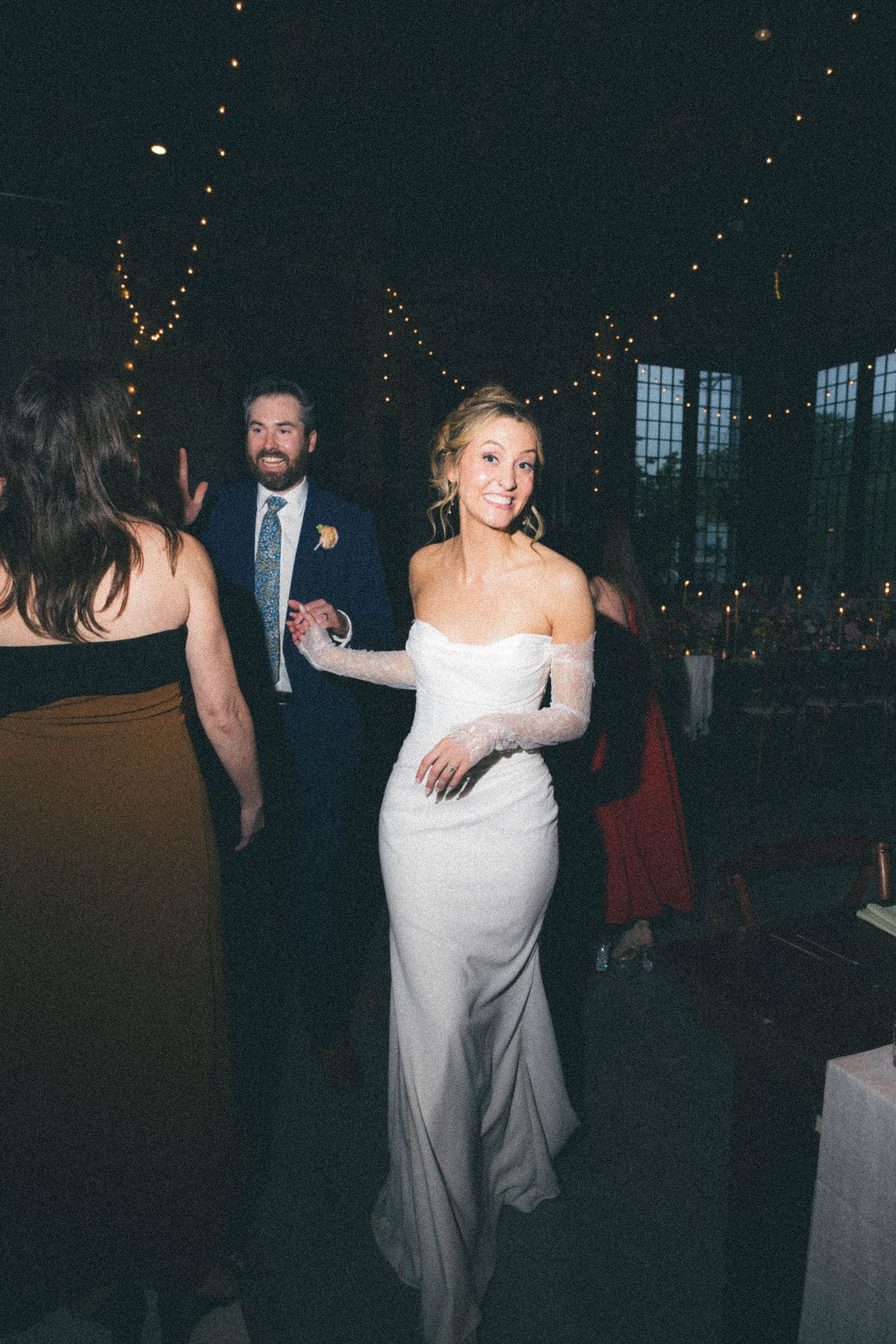 Bride in elegant off-shoulder white gown walks through wedding reception with string lights.