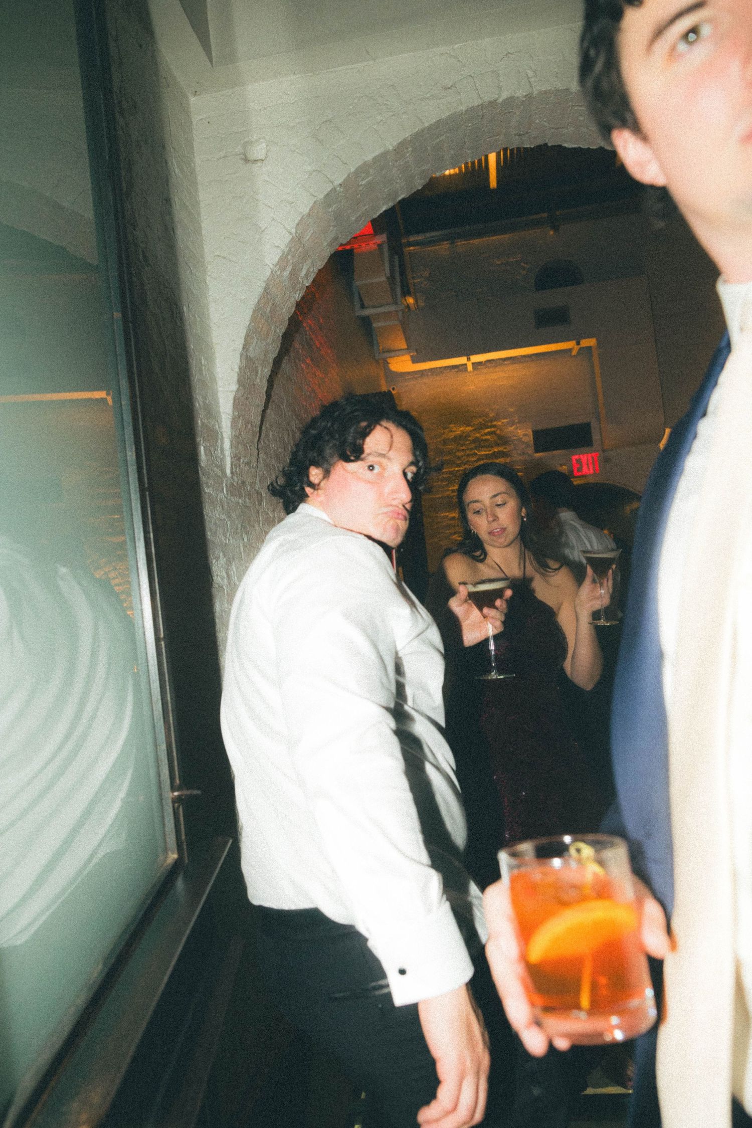 Guests in formal attire socialize with cocktails near an arched stone doorway at a wedding.