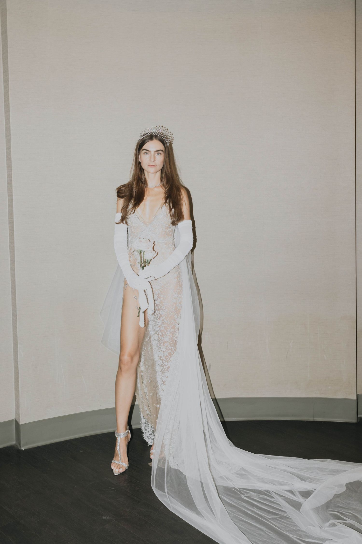 Bride in sheer lace wedding gown with high slit, crystal crown, and long white gloves with flowing veil.