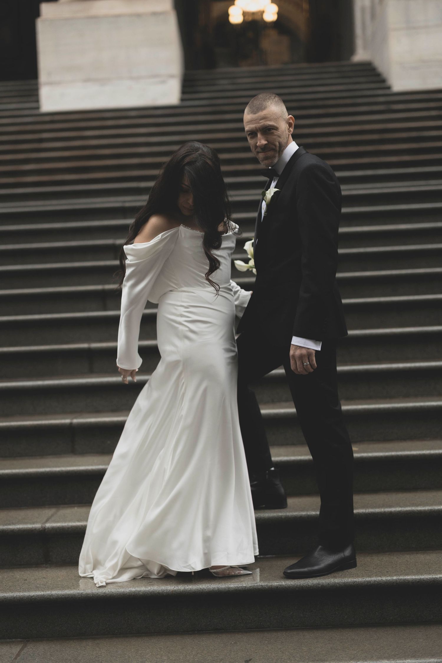 Bride and groom in elegant wedding attire walking down grand stone staircase together.