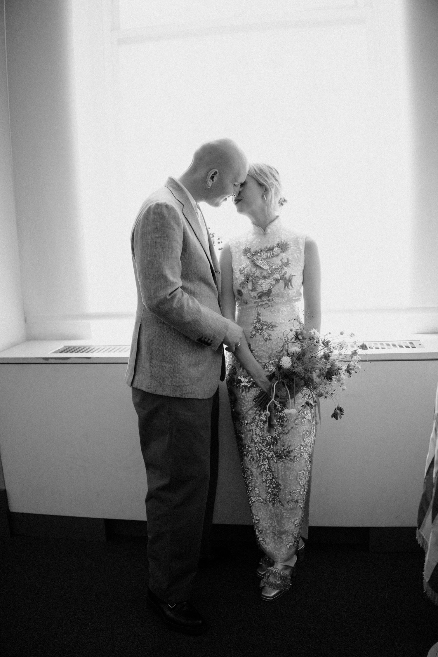 Black and white romantic portrait of bride and groom touching foreheads by bright window.