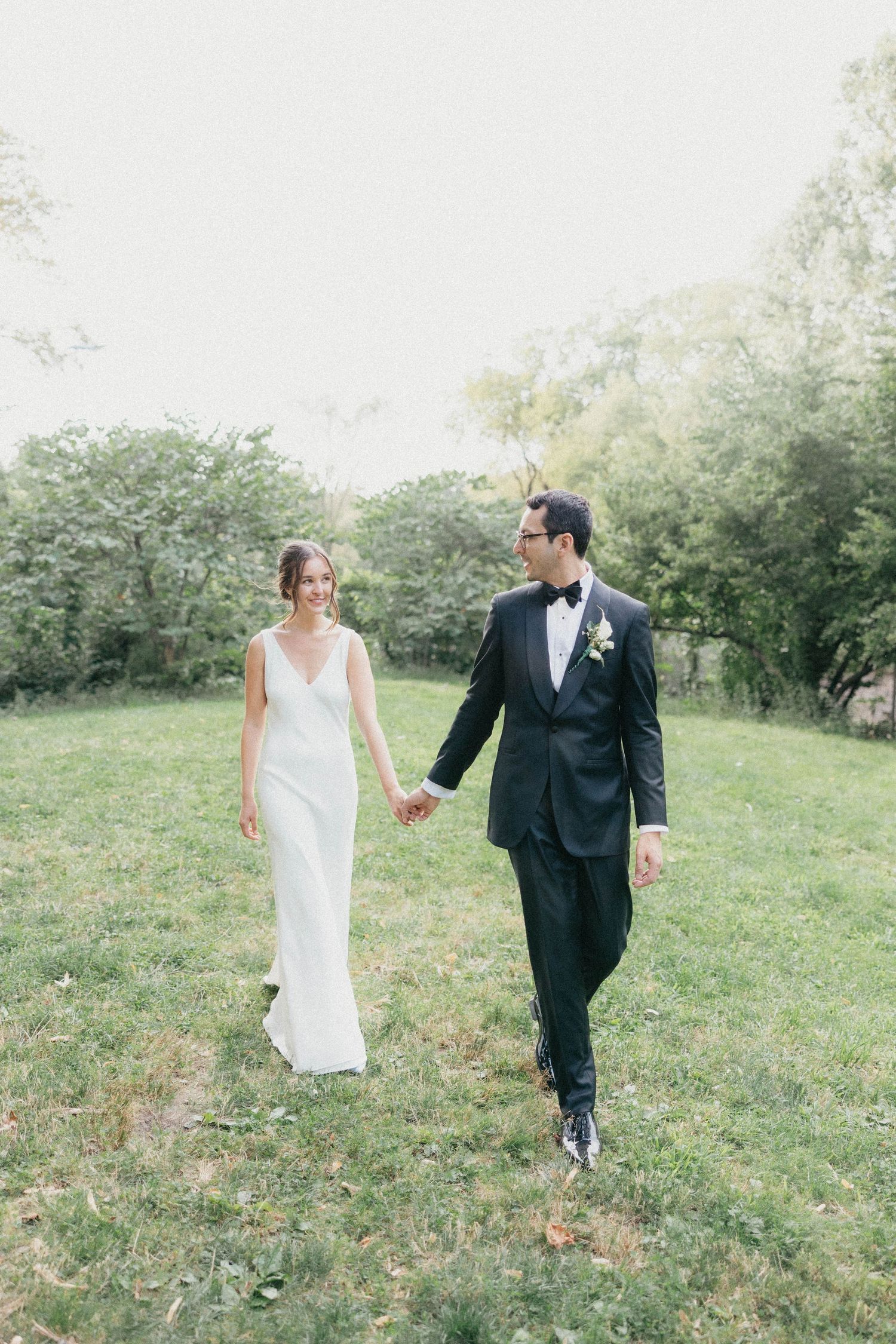 Smiling bride in white gown and groom in black tuxedo holding hands walking across green lawn.