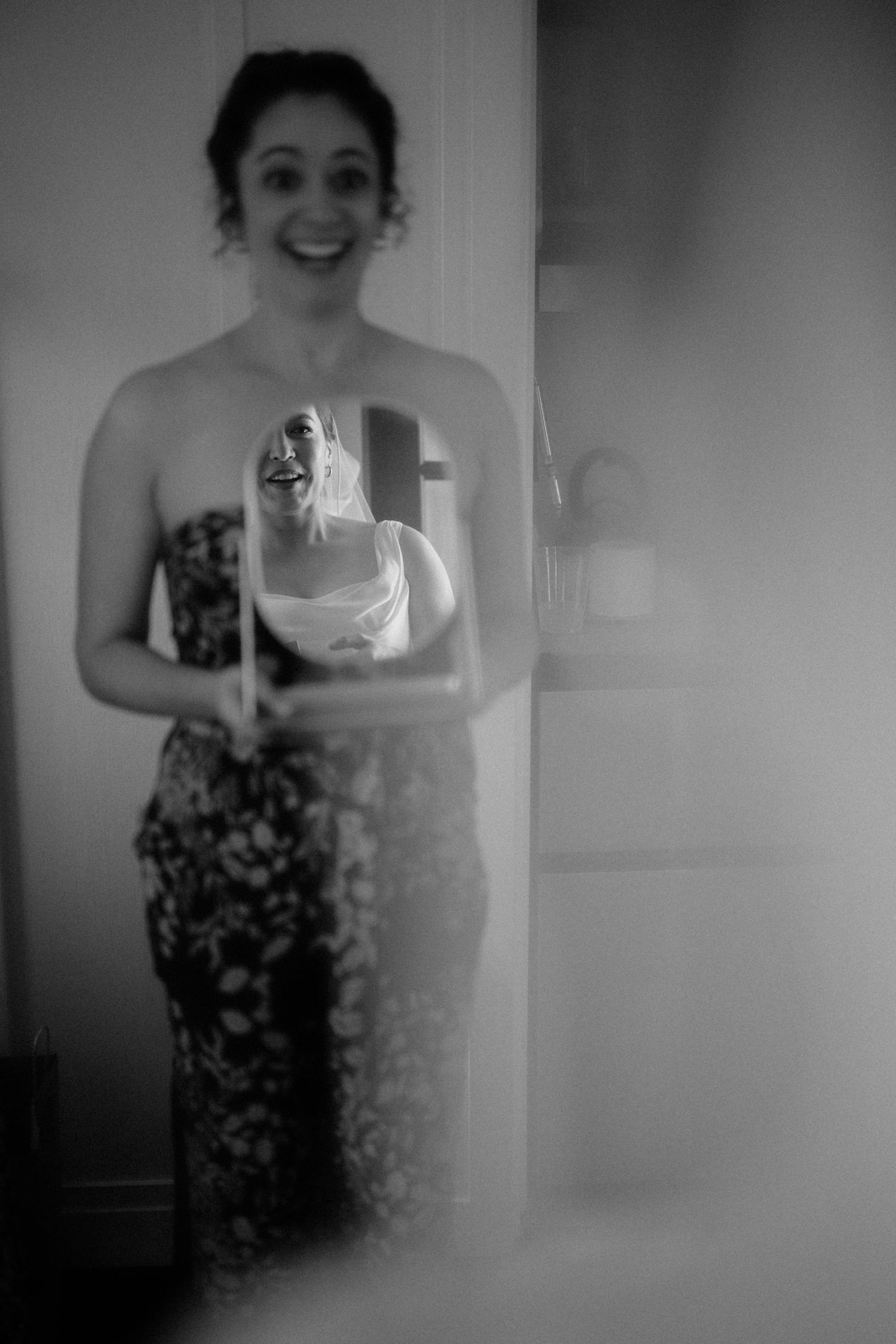 Smiling bridesmaid in floral dress holds mirror reflecting bride in black and white photo.