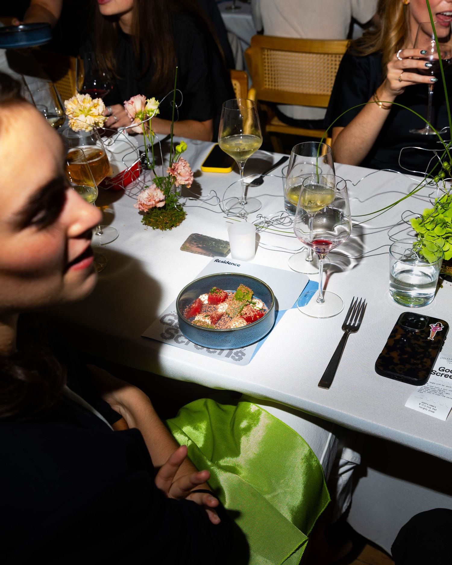 Guests dine at a long white table set with wine glasses, a blue bowl of colorful appetizer, pink flowers and green foliage.