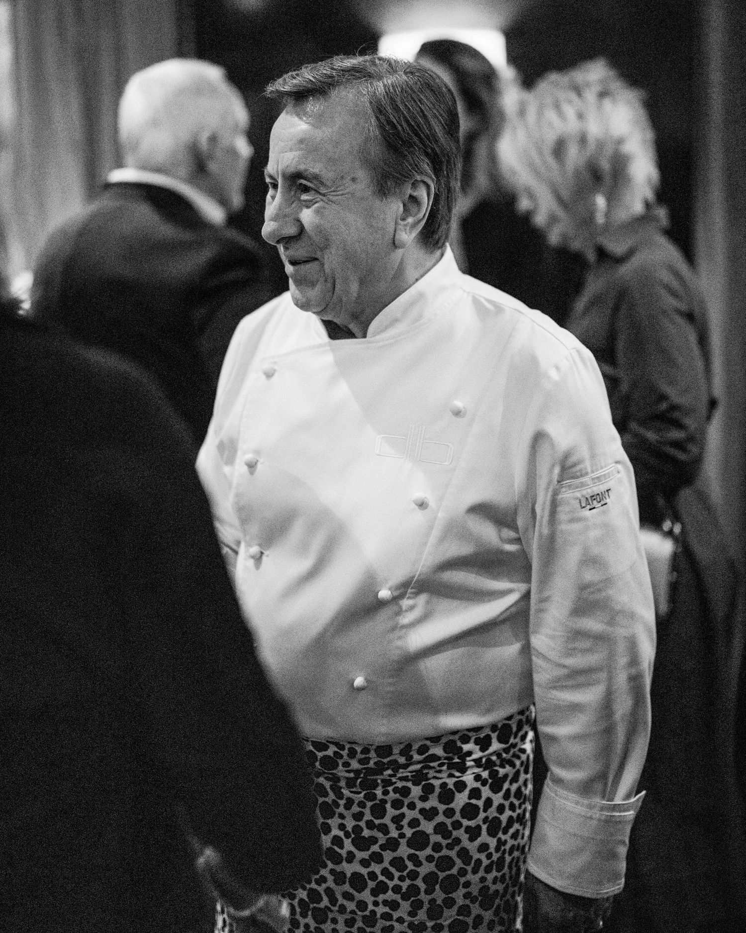 Smiling chef in a white double-breasted jacket and leopard print apron stands among guests in a black and white photo.