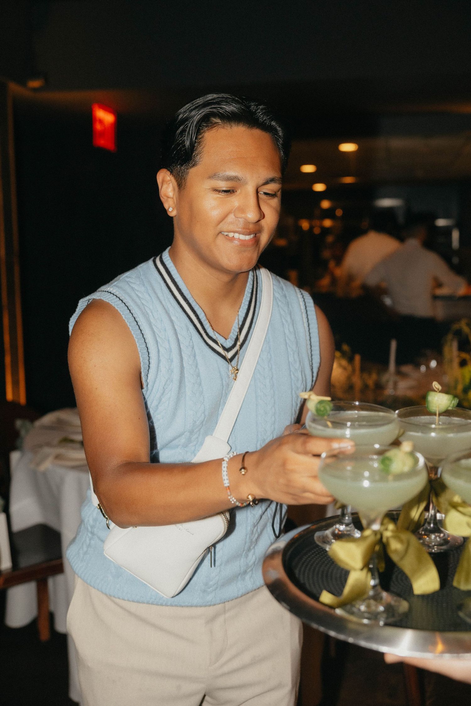 Smiling man in a light blue sweater vest holds a coupe cocktail garnished with lime from a tray of drinks with yellow ribbons.