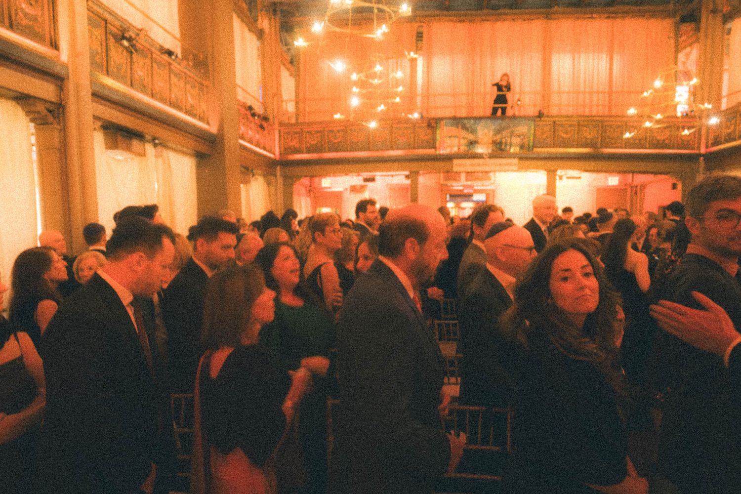 Warm-toned wide shot of wedding reception guests mingling in grand ballroom with ornate architecture.