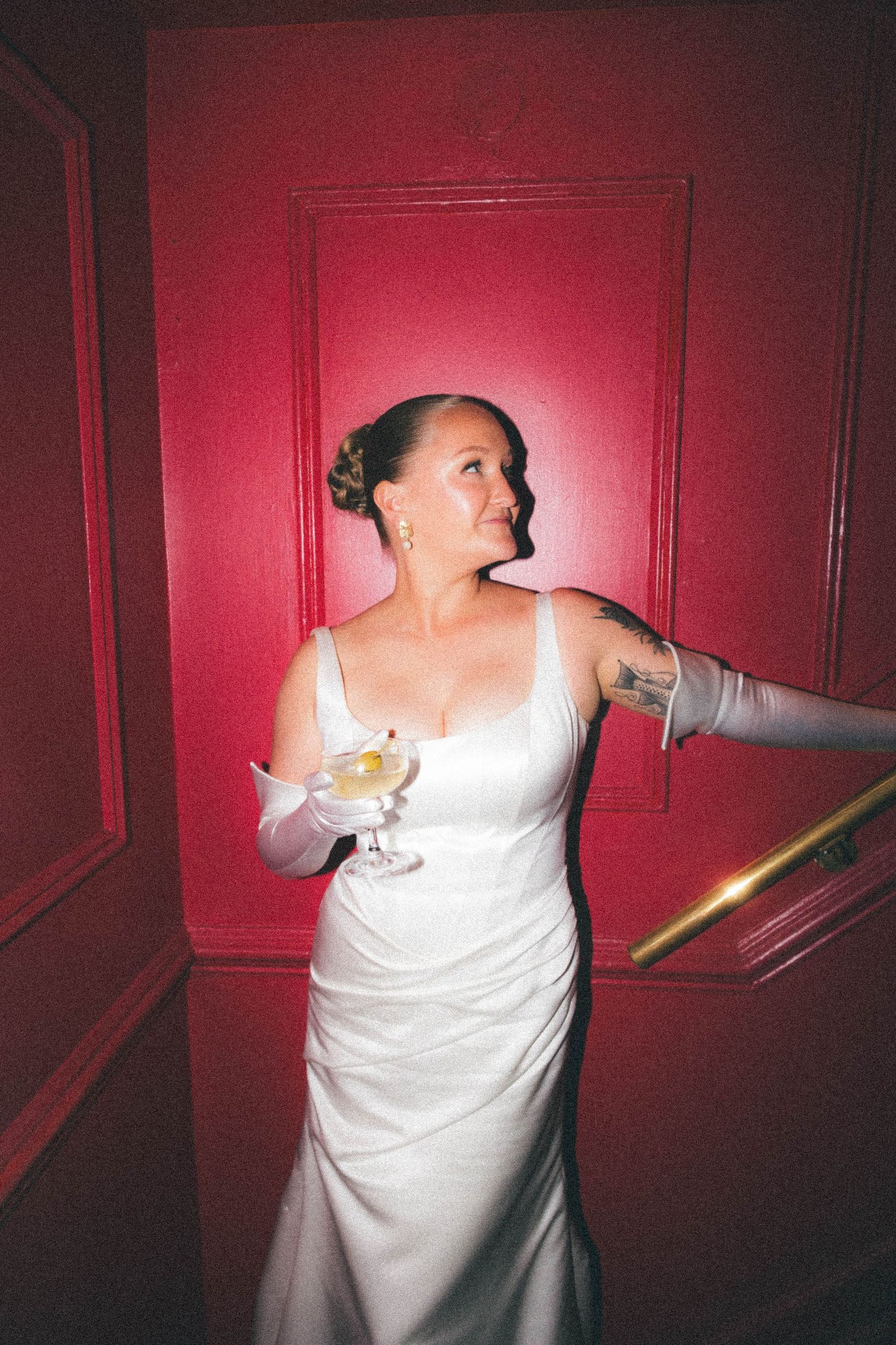 Bride in white gown holding martini glass while leaning on gold railing against red wall.