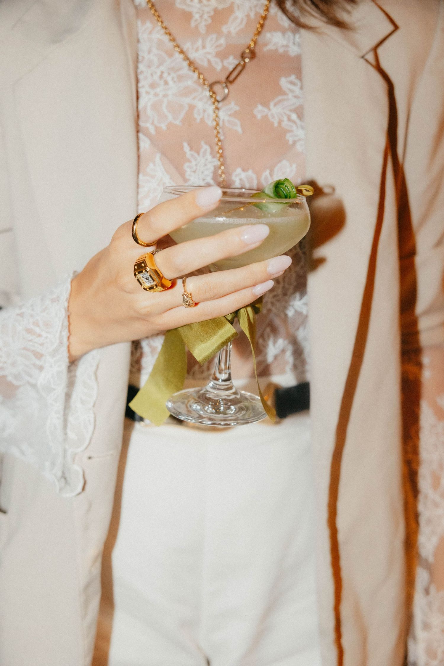 Woman wearing gold rings and a layered necklace holds a pale green coupe cocktail with a garnish, dressed in a cream blazer and lace top.