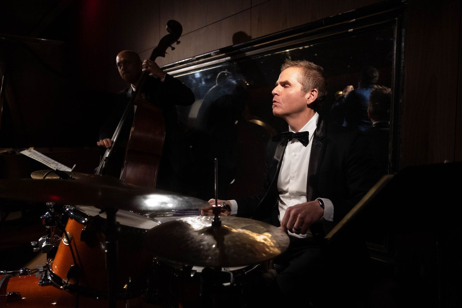 Jazz drummer in a tuxedo and bow tie plays a drum kit with cymbals while an upright bass player performs behind him in a dimly lit club.