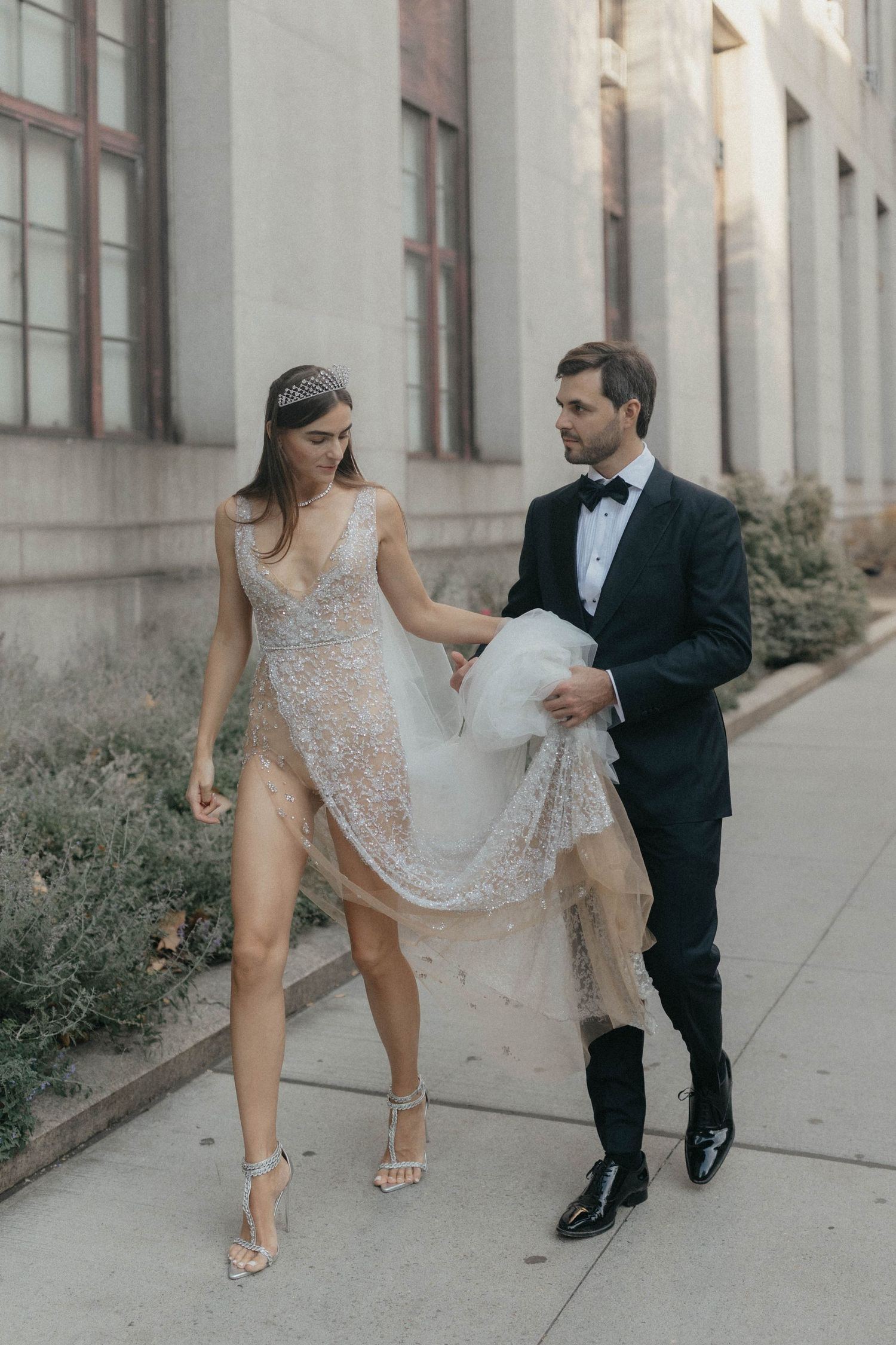 Groom in tuxedo carries bride's beaded lace wedding gown train as couple walks along urban sidewalk.