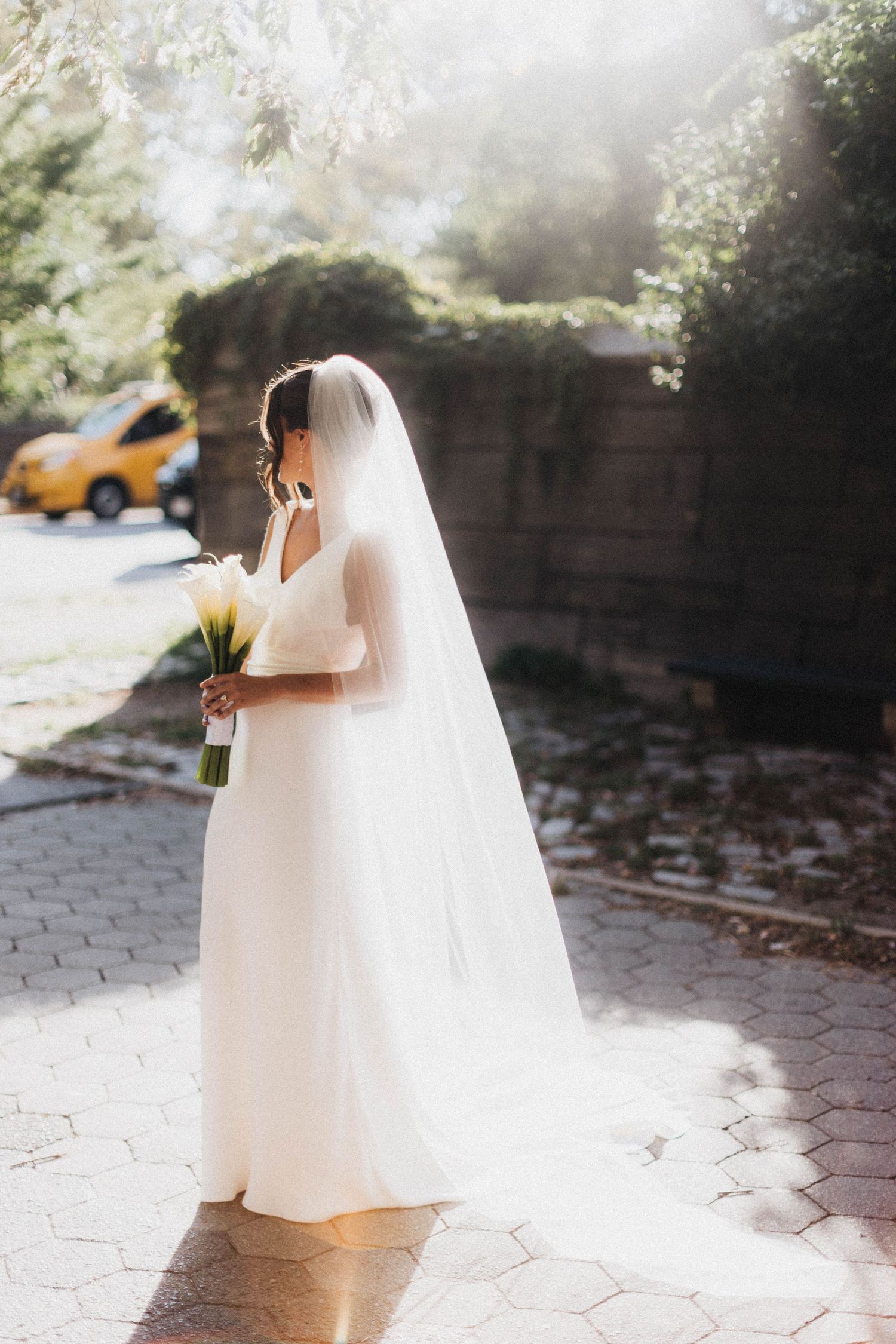 Bride in white wedding gown with long veil holding calla lily bouquet on sunlit cobblestone street.