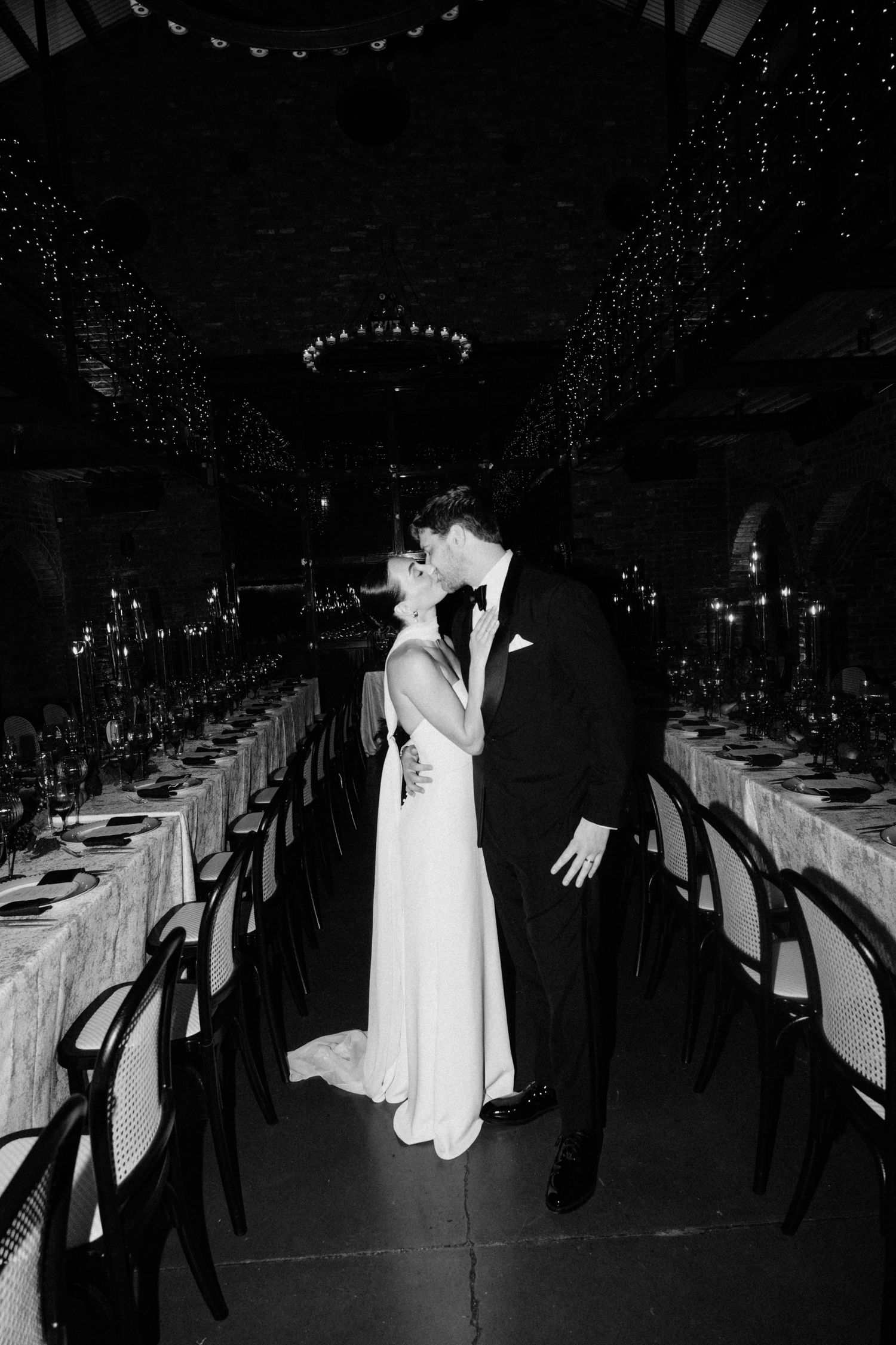 Bride and groom kissing between long reception tables with twinkling lights overhead.