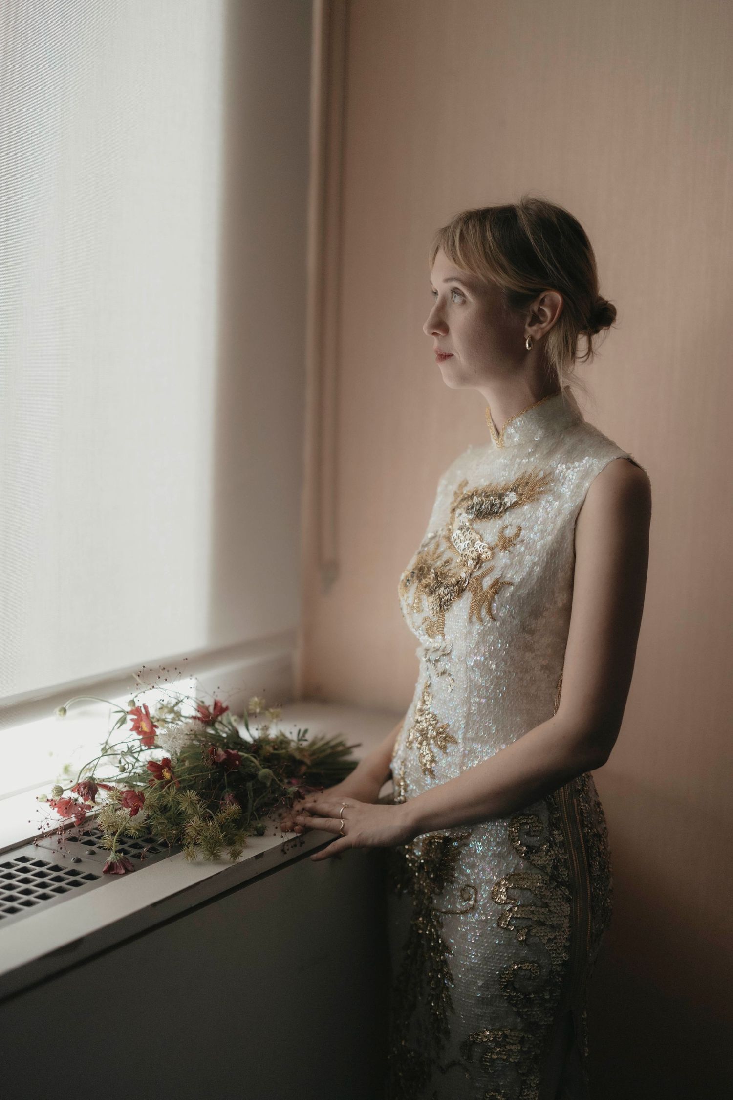 Elegant bride in embroidered gown holding floral bouquet by a window in soft natural light.
