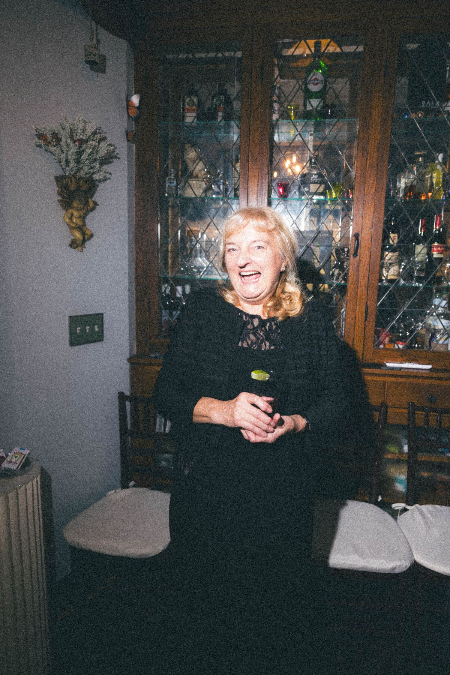 Laughing blonde woman in black lace dress standing near vintage wooden bar cabinet at celebration.