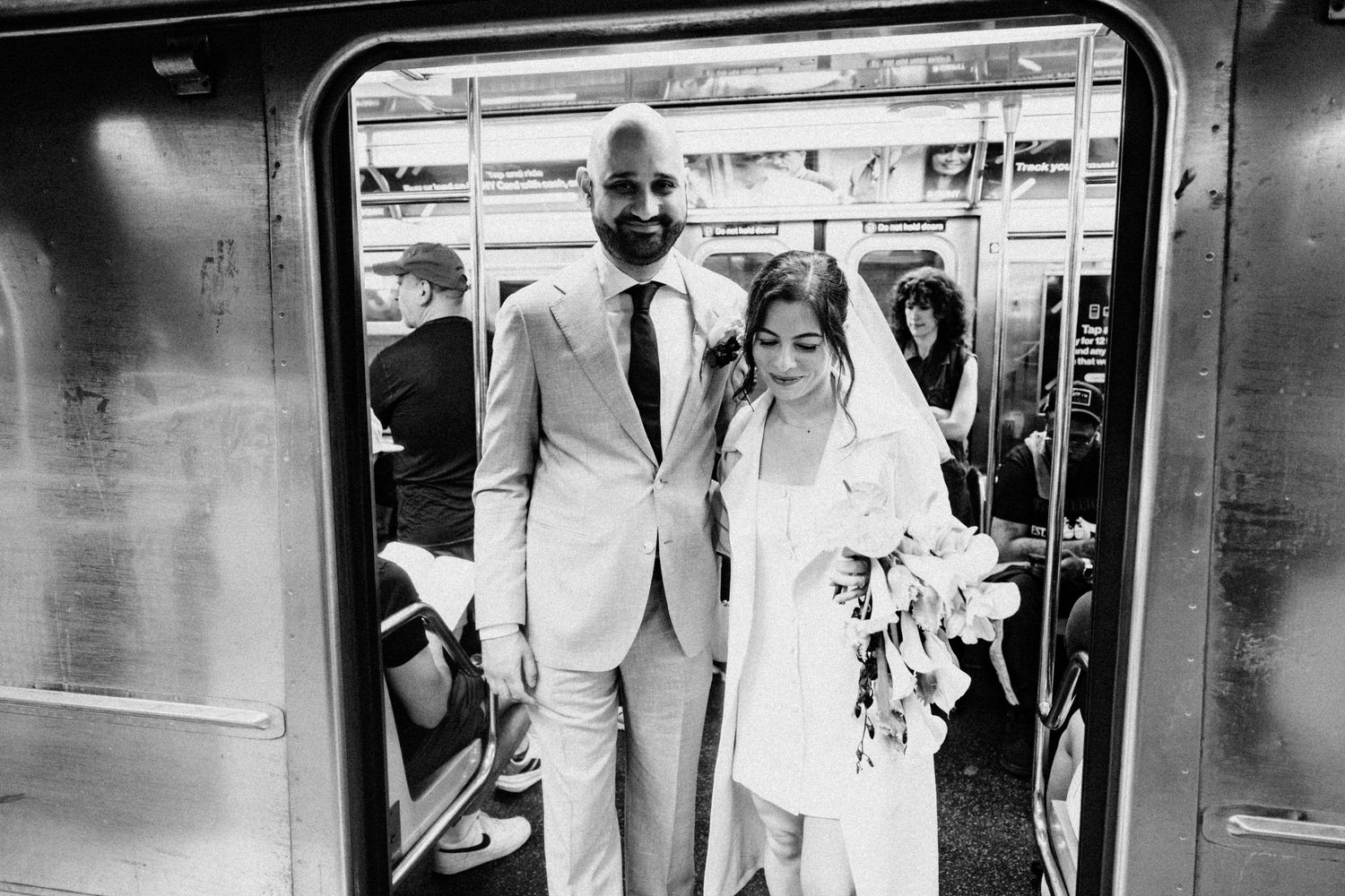Black and white photo of a bride and groom standing in subway train doors holding a bouquet.