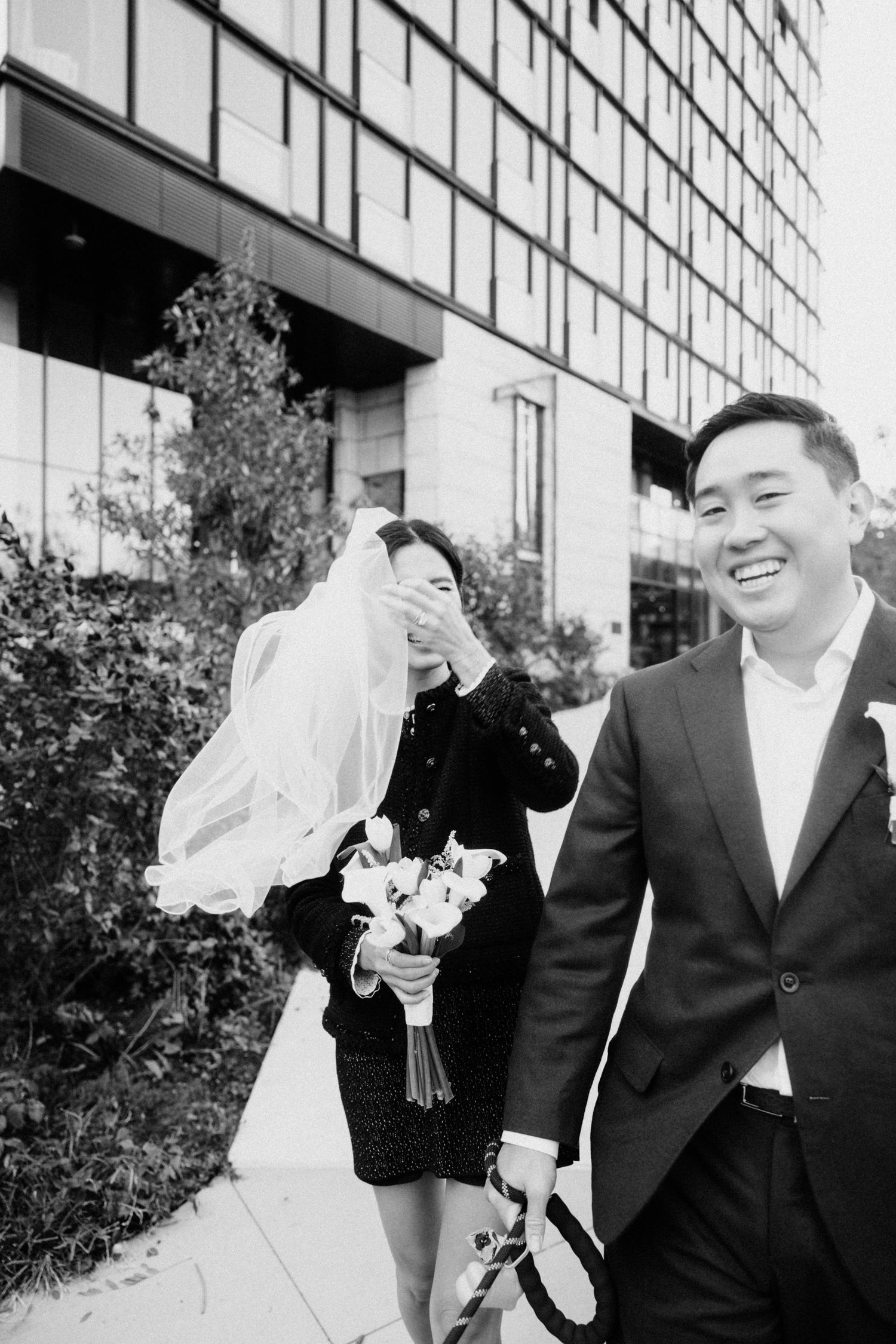 Black and white photo of smiling groom walking with bride covering face with veil.