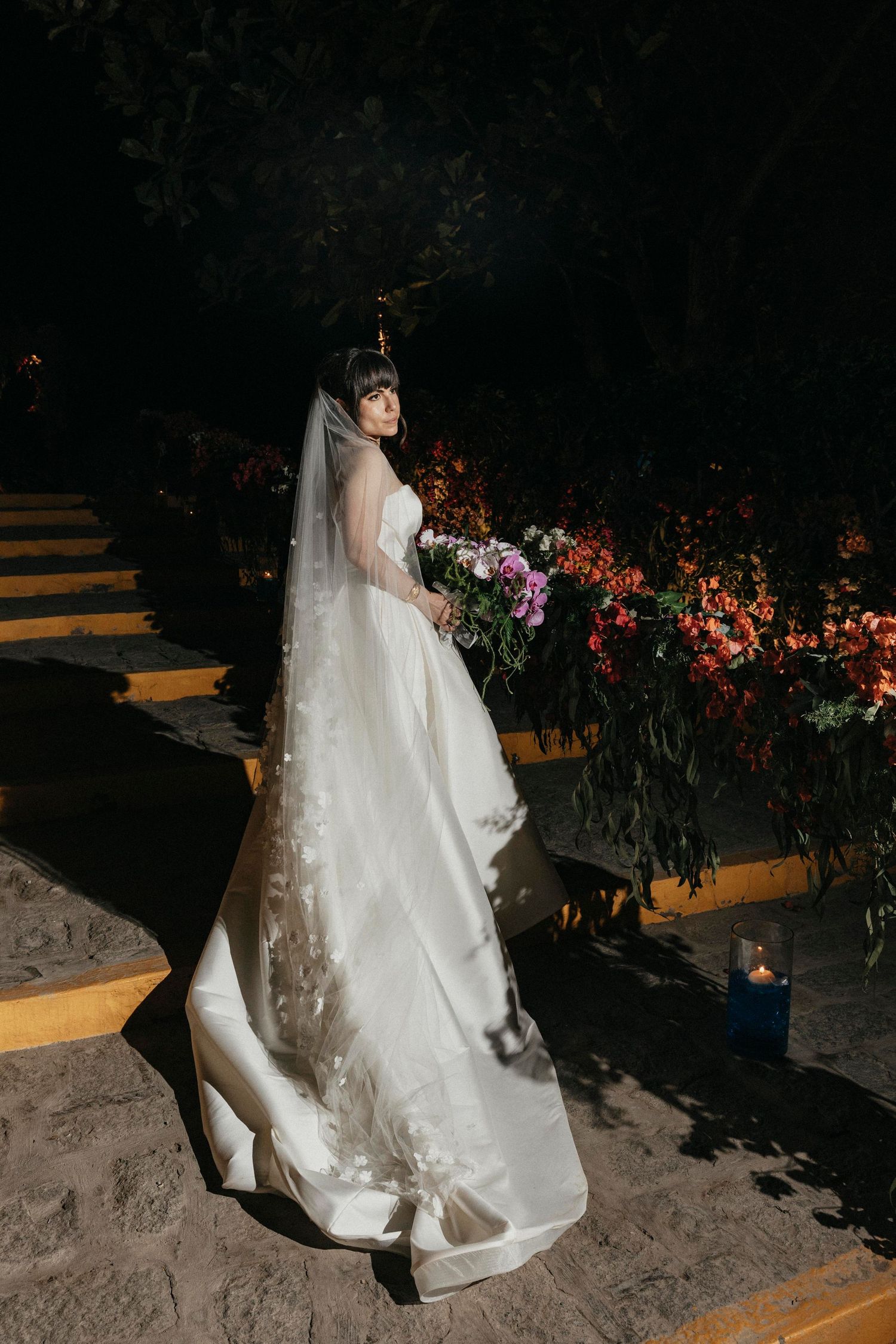 Elegant bride in white gown with floral veil poses near colorful floral arrangements at night wedding.