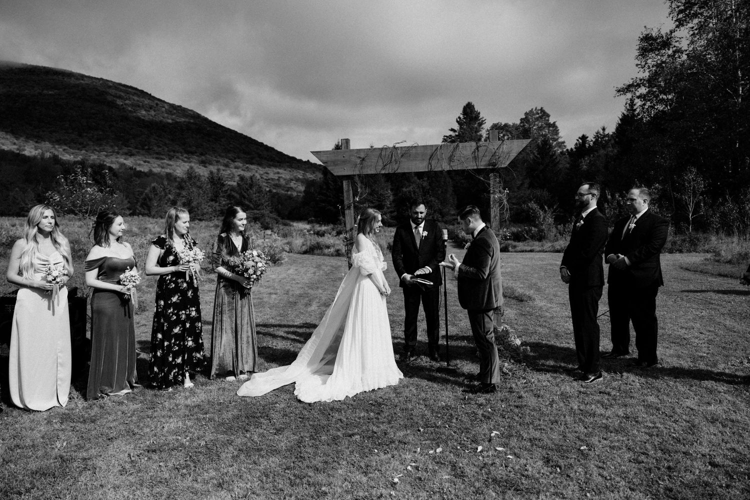 Black and white photo of wedding ceremony with bridal party at outdoor rustic altar.