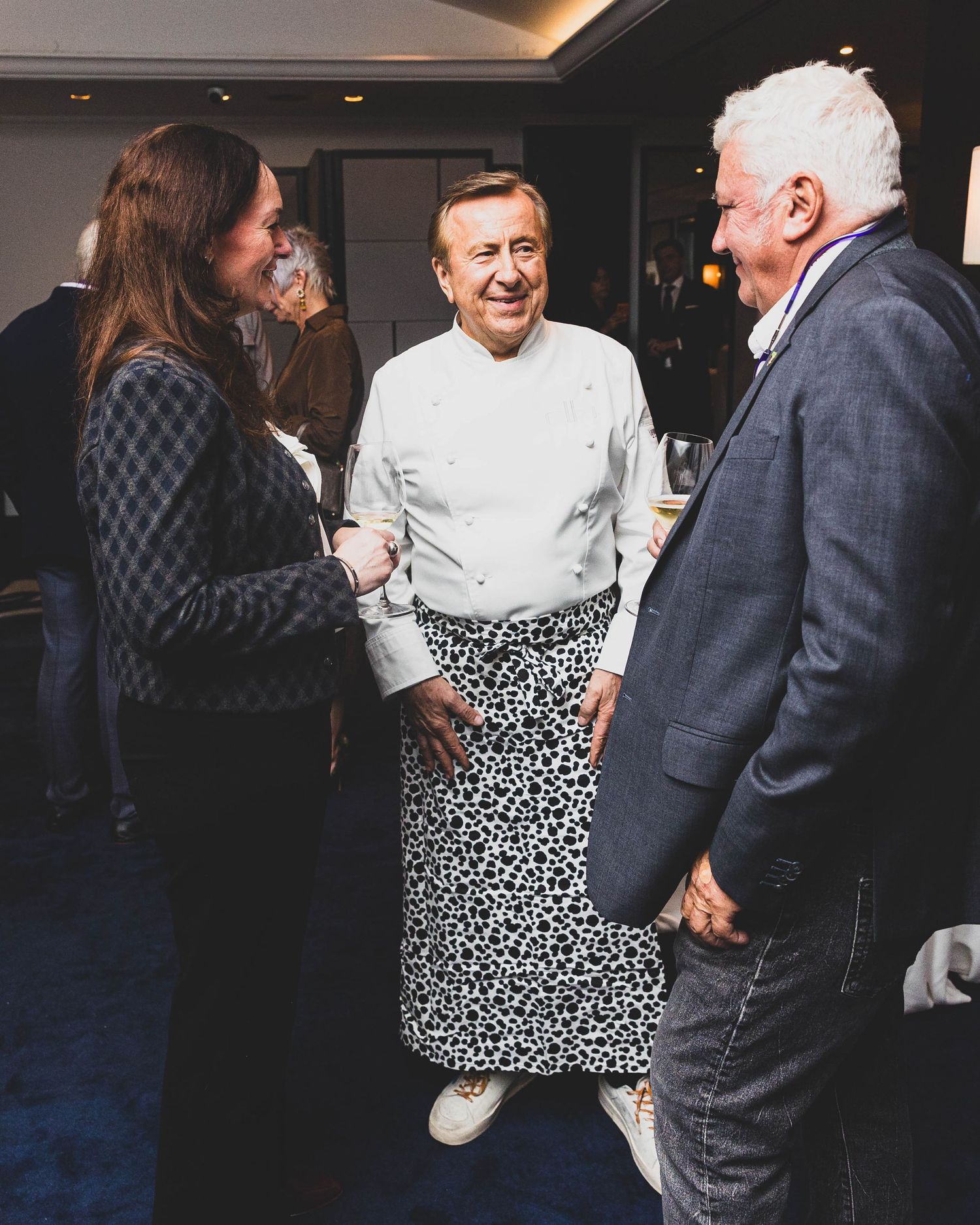 Smiling chef in a white jacket and spotted apron chats with a woman in a tweed jacket and a gray-haired man in a blazer at an indoor event.