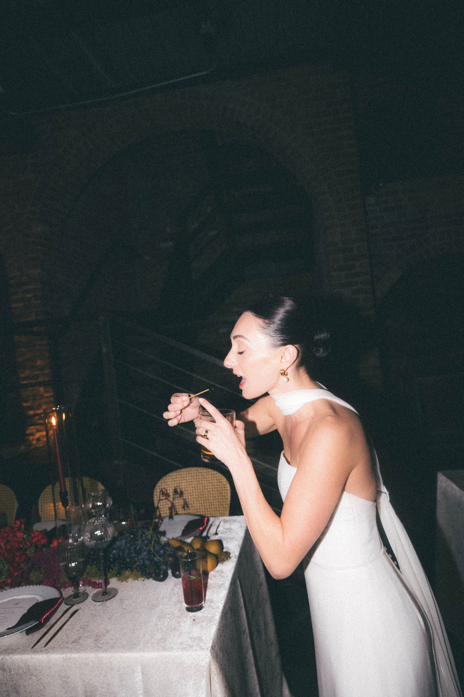 Bride in white halter gown eating at candlelit wedding reception table in rustic brick venue.