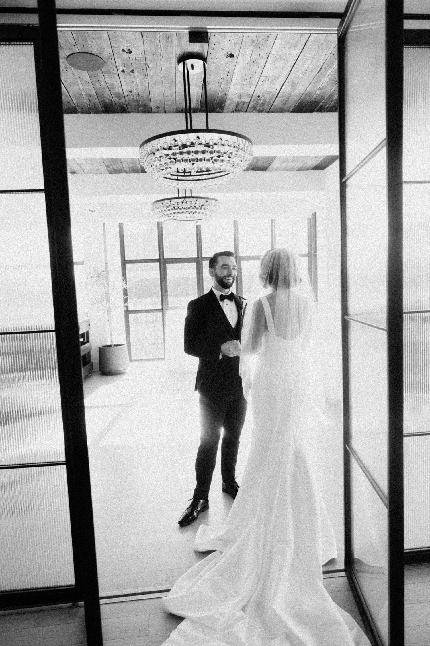 Bride and groom sharing first look moment in bright room with crystal chandelier in black and white photo.