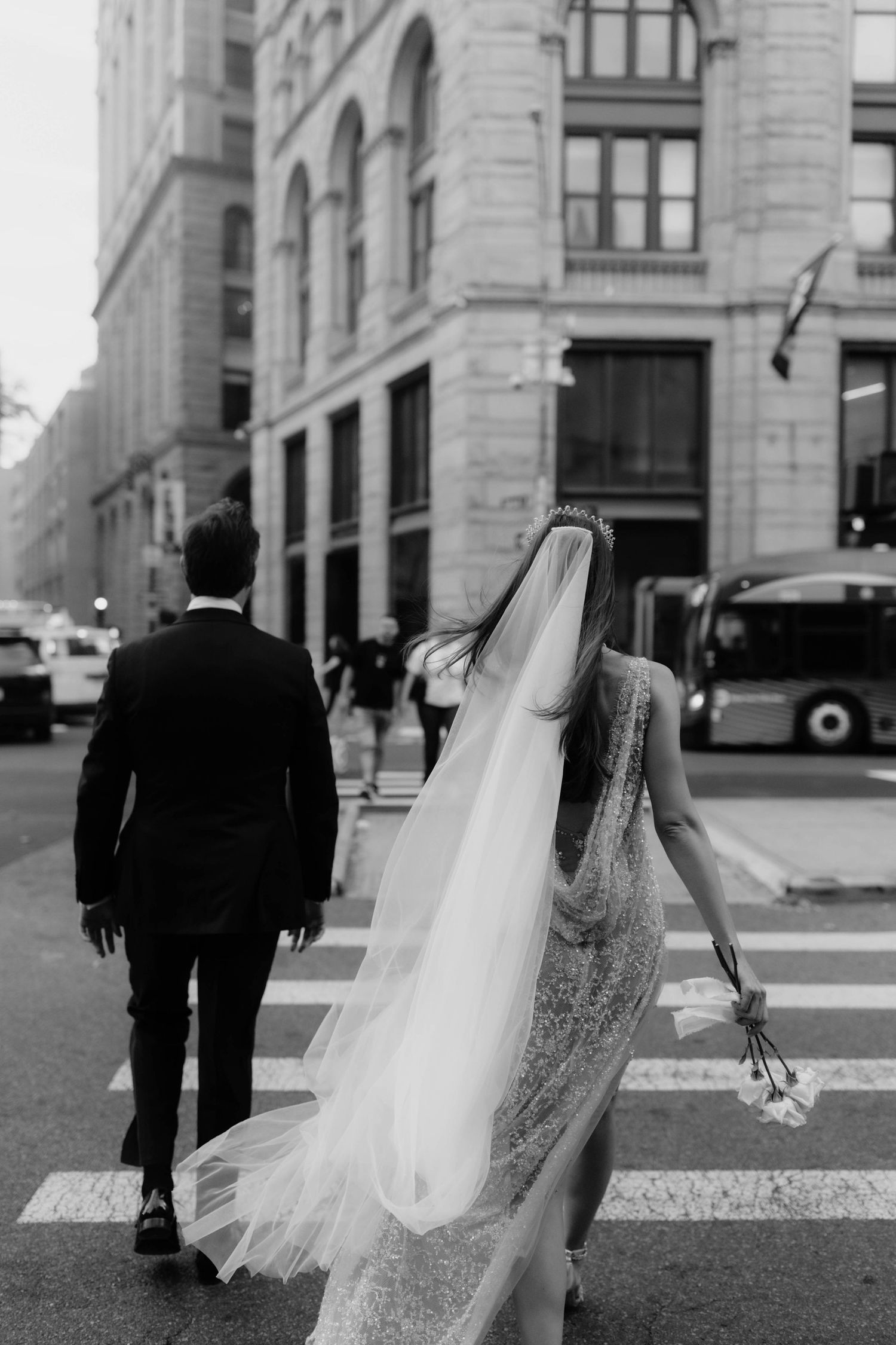 Black and white photo of bride and groom crossing a city street, her long veil and bouquet flowing in the wind.