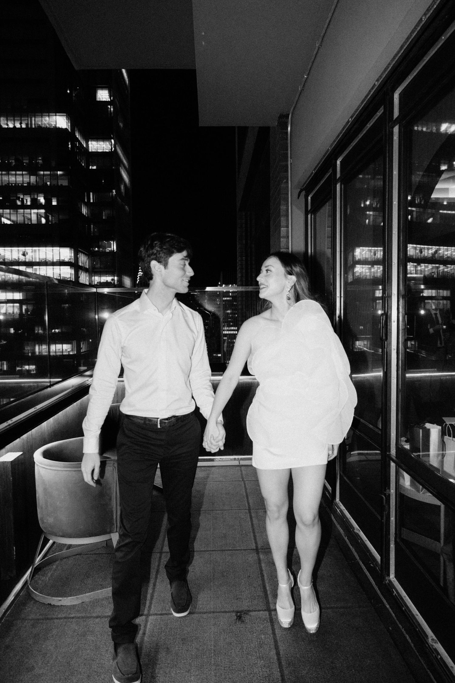 Black and white photo of couple holding hands walking on outdoor terrace with city lights at night.