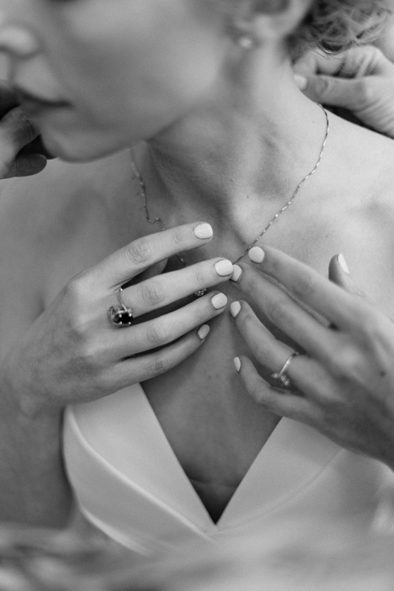 Close-up black and white photo of bride having delicate necklace fastened while wearing engagement and wedding rings.