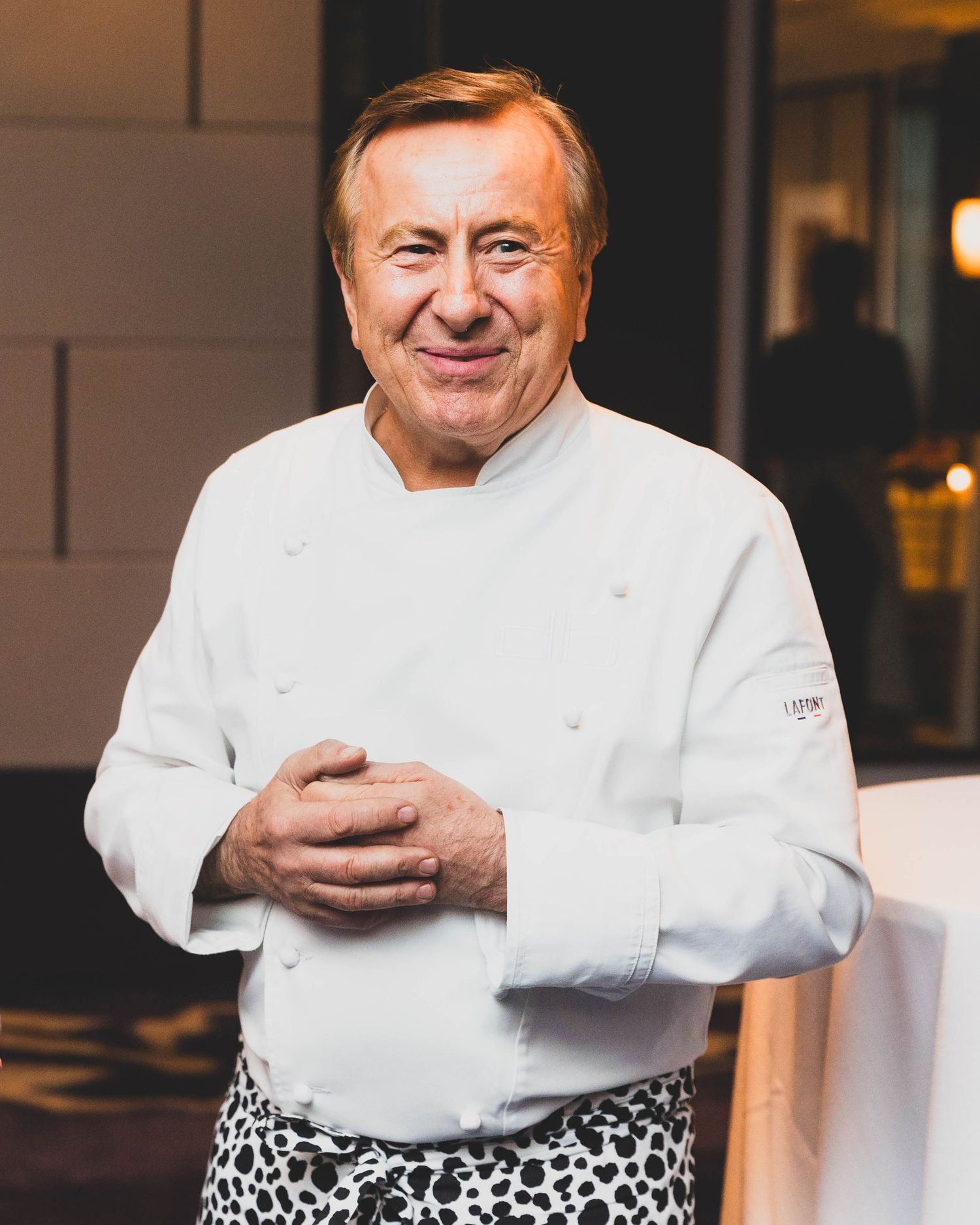 Smiling older chef in a white double-breasted chef jacket and spotted apron stands with hands clasped in an elegant restaurant dining room.