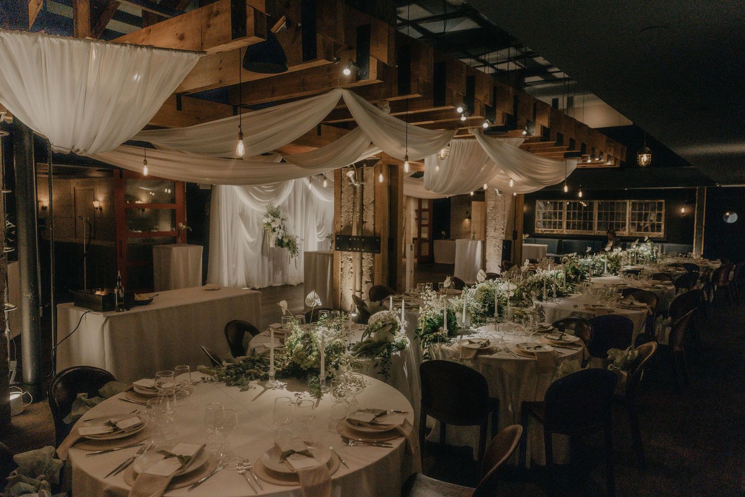Elegant wedding reception venue with draped white fabric, warm string lights, and candlelit tables set with greenery centerpieces.