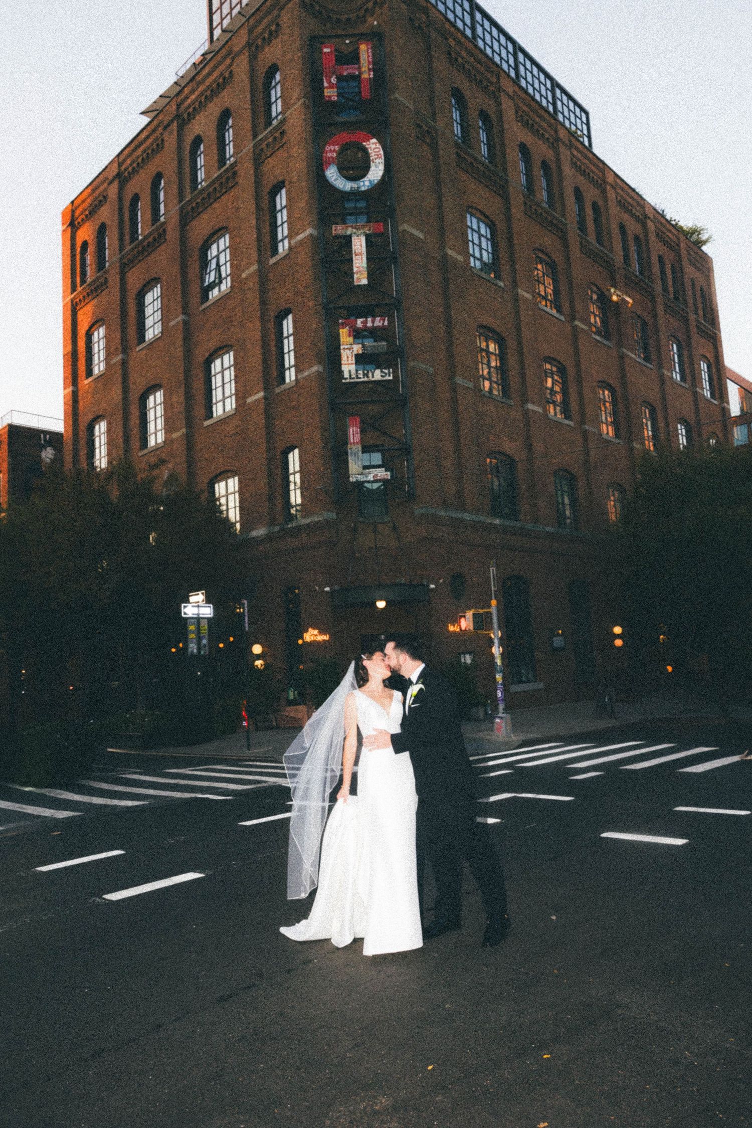 Bride and groom embrace at dusk on a city street corner in front of a brick hotel building.