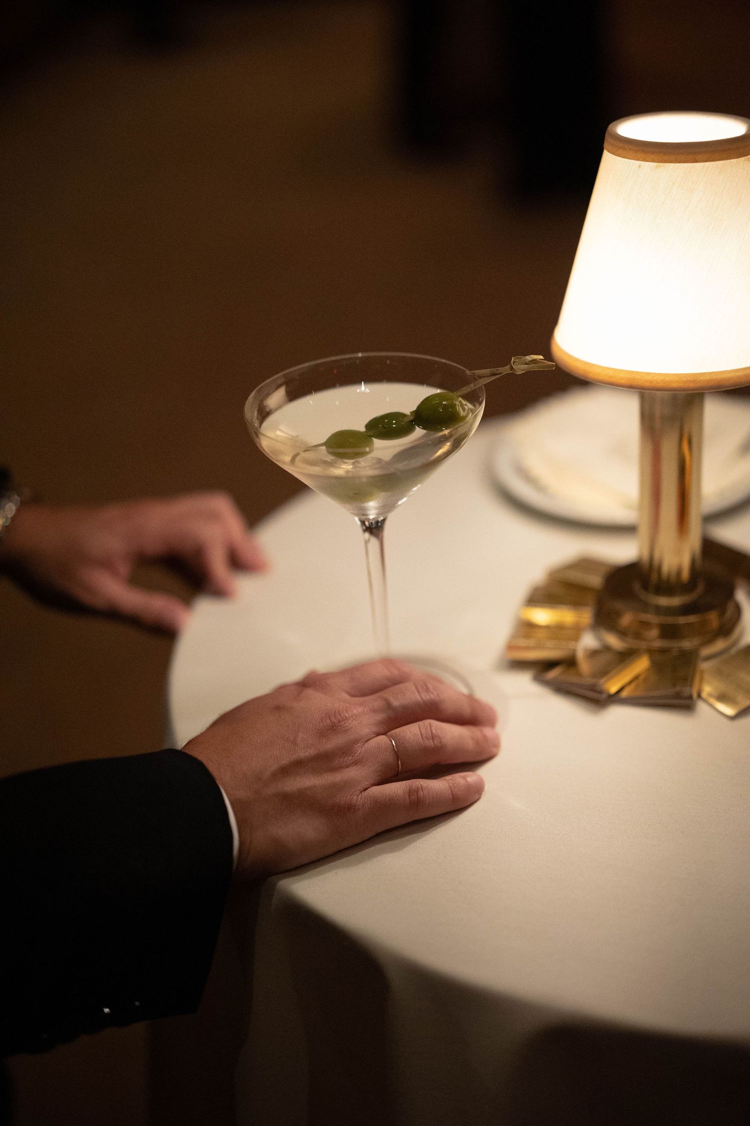 A martini with three green olives on a pick sits on a white clothed table beside a glowing brass lamp as two hands rest nearby.