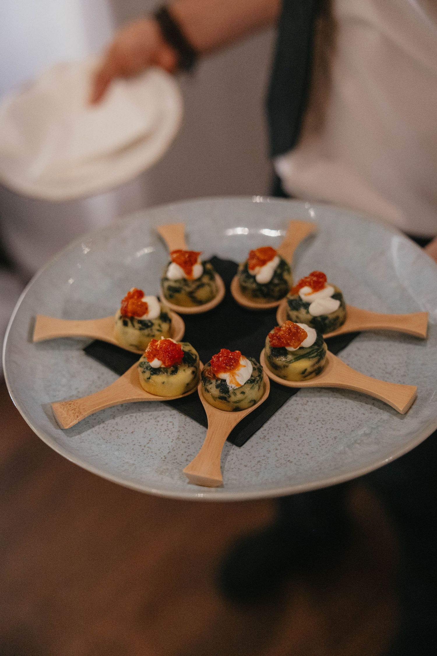 A server holds a gray plate of spinach mini quiche appetizers topped with cream and red caviar, served on wooden spoons.