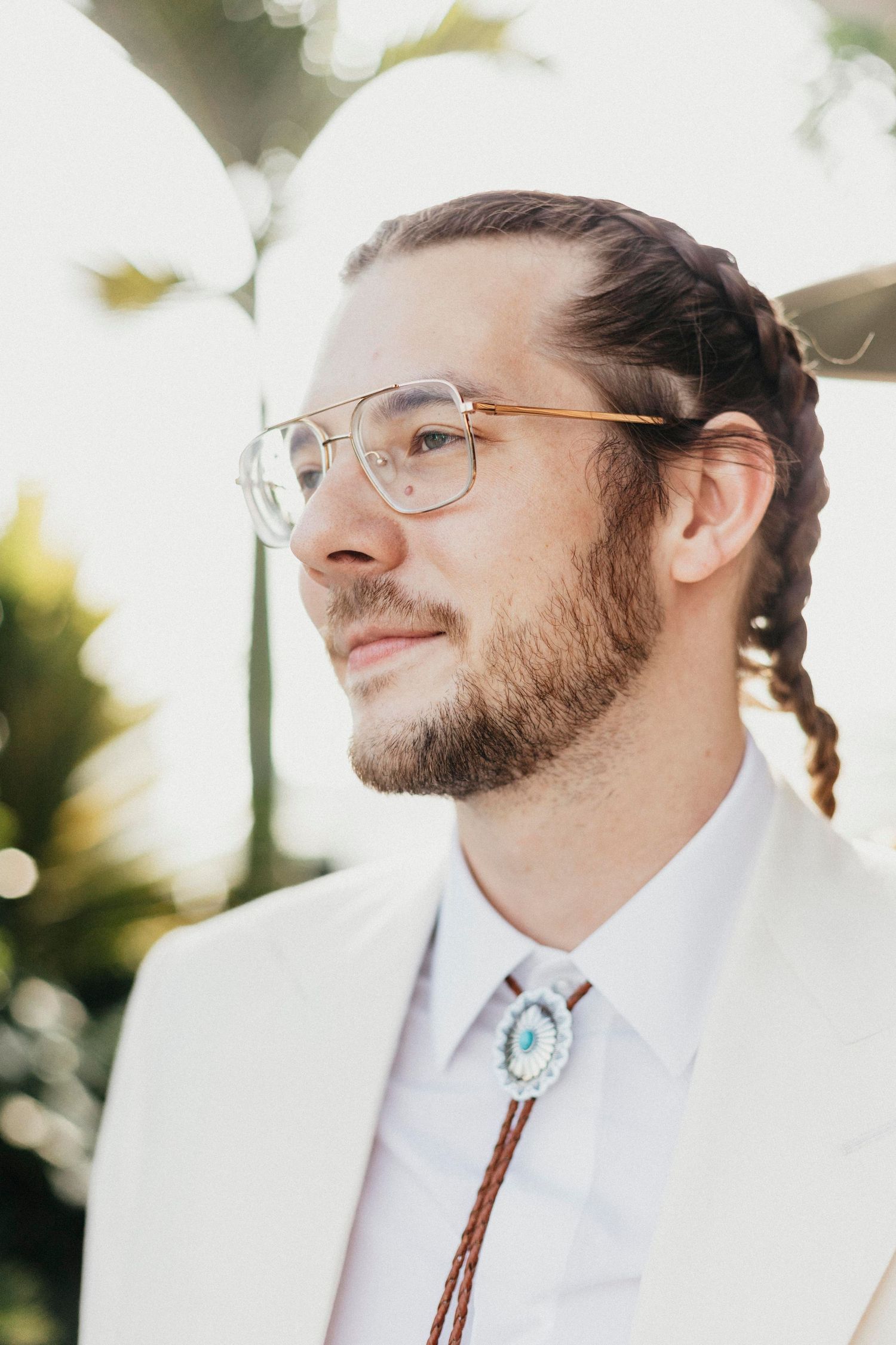 Portrait of groom with braided hair wearing glasses and bolo tie in white wedding suit.