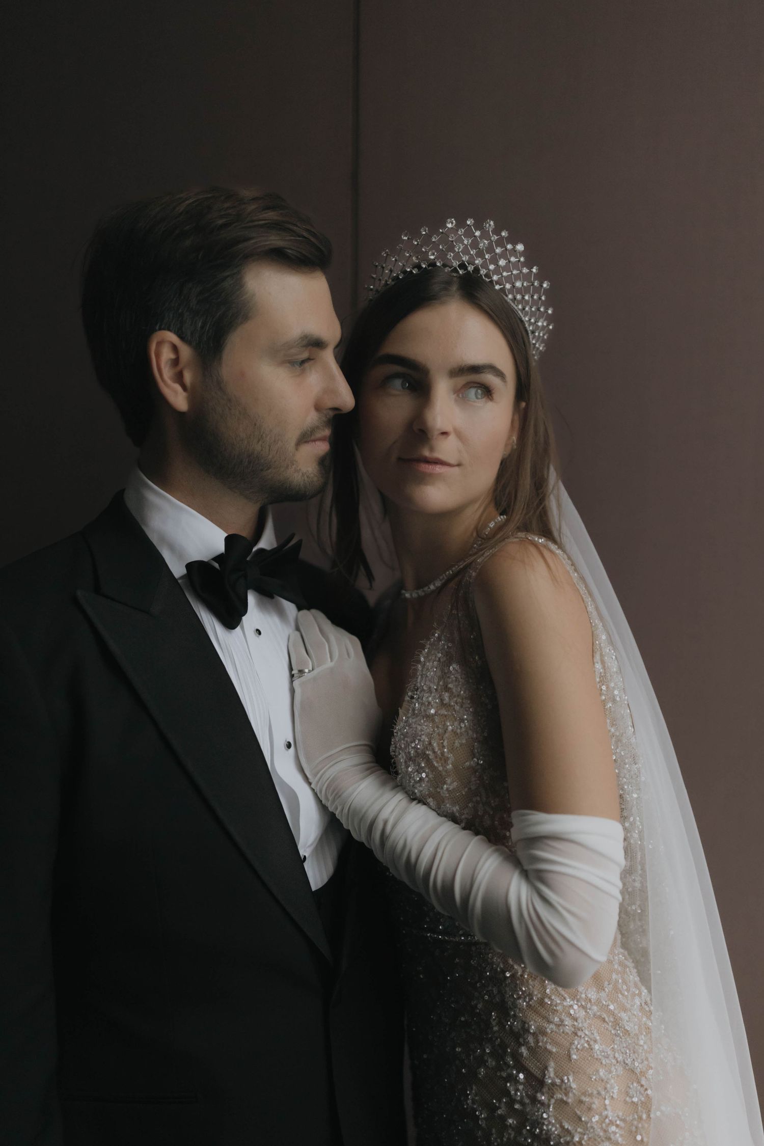 Elegant bride in beaded wedding gown with crystal headpiece and white gloves posing with dapper groom.