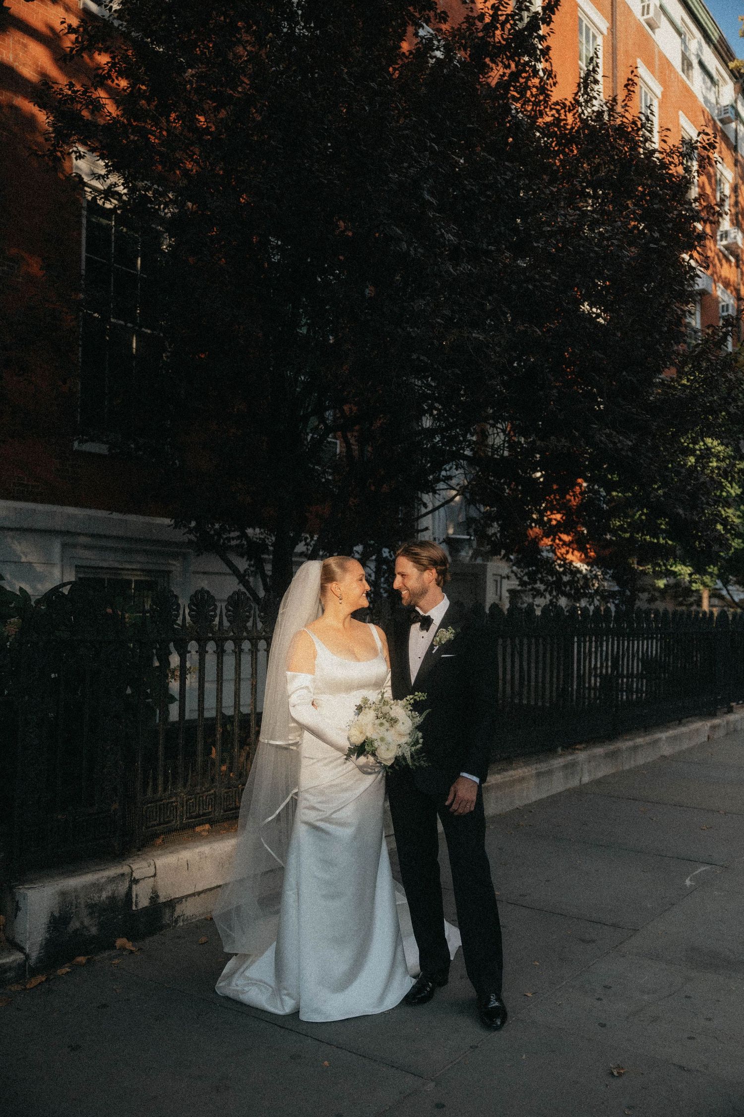 Bride in off-shoulder gown with veil and groom in tuxedo share romantic moment on sunlit city sidewalk.