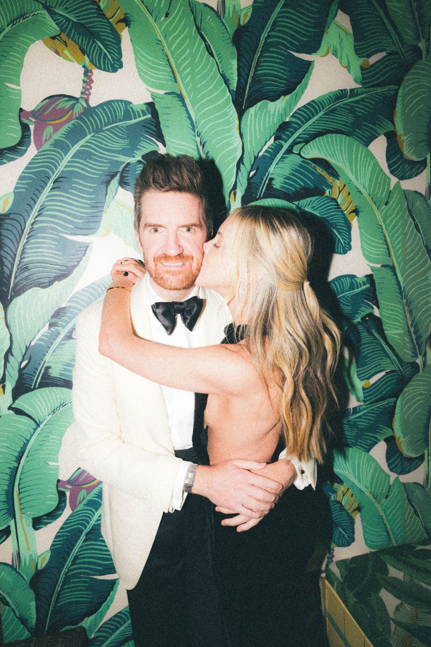 Couple kissing and embracing in front of tropical banana leaf wallpaper at wedding.