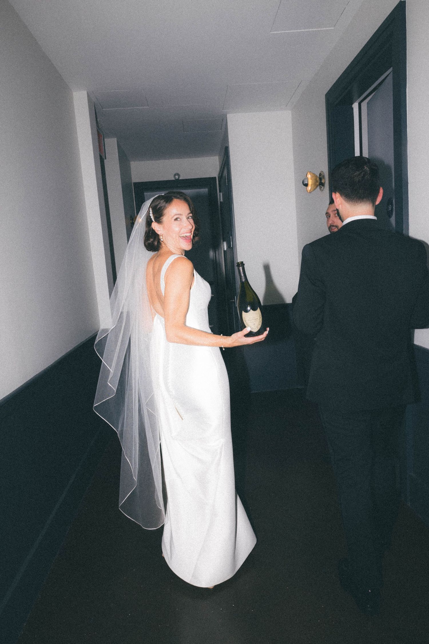 Smiling bride in white gown and veil holding champagne bottle while walking hotel corridor with groom.