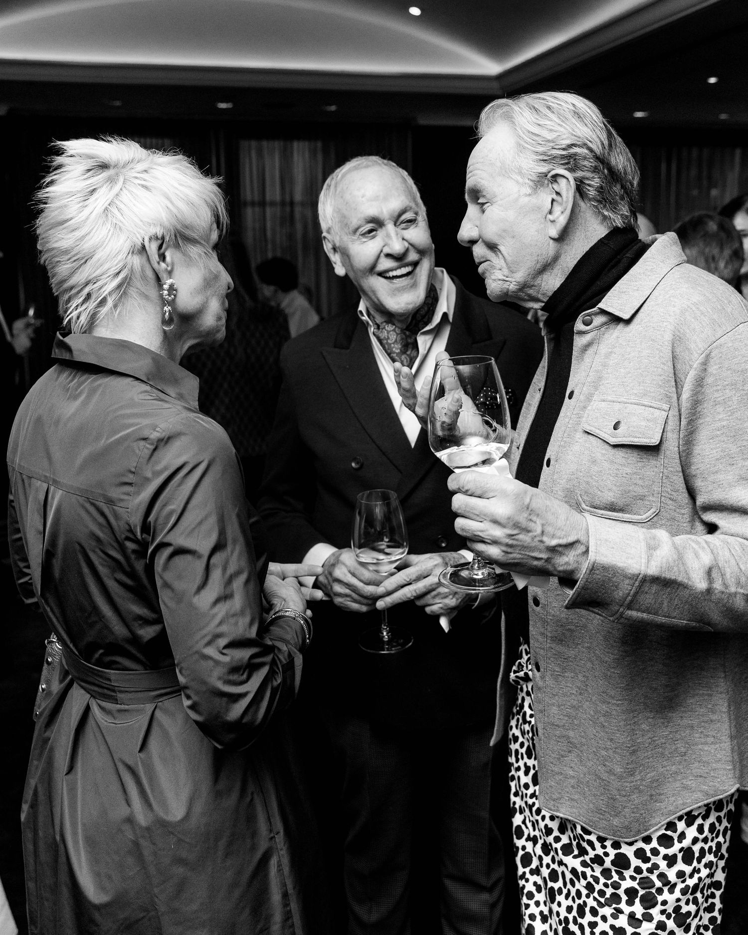 Three smiling guests chat and hold wine glasses at an elegant evening reception in a dimly lit room, shown in black and white.