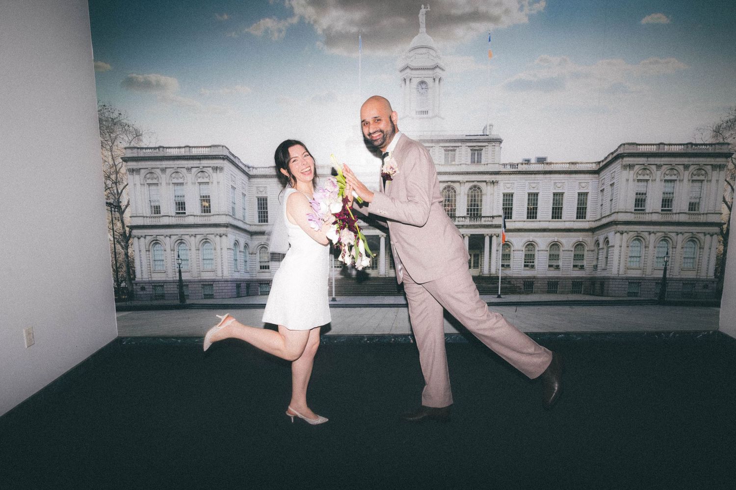 Joyful bride and groom posing playfully in front of a city hall backdrop with bouquet.